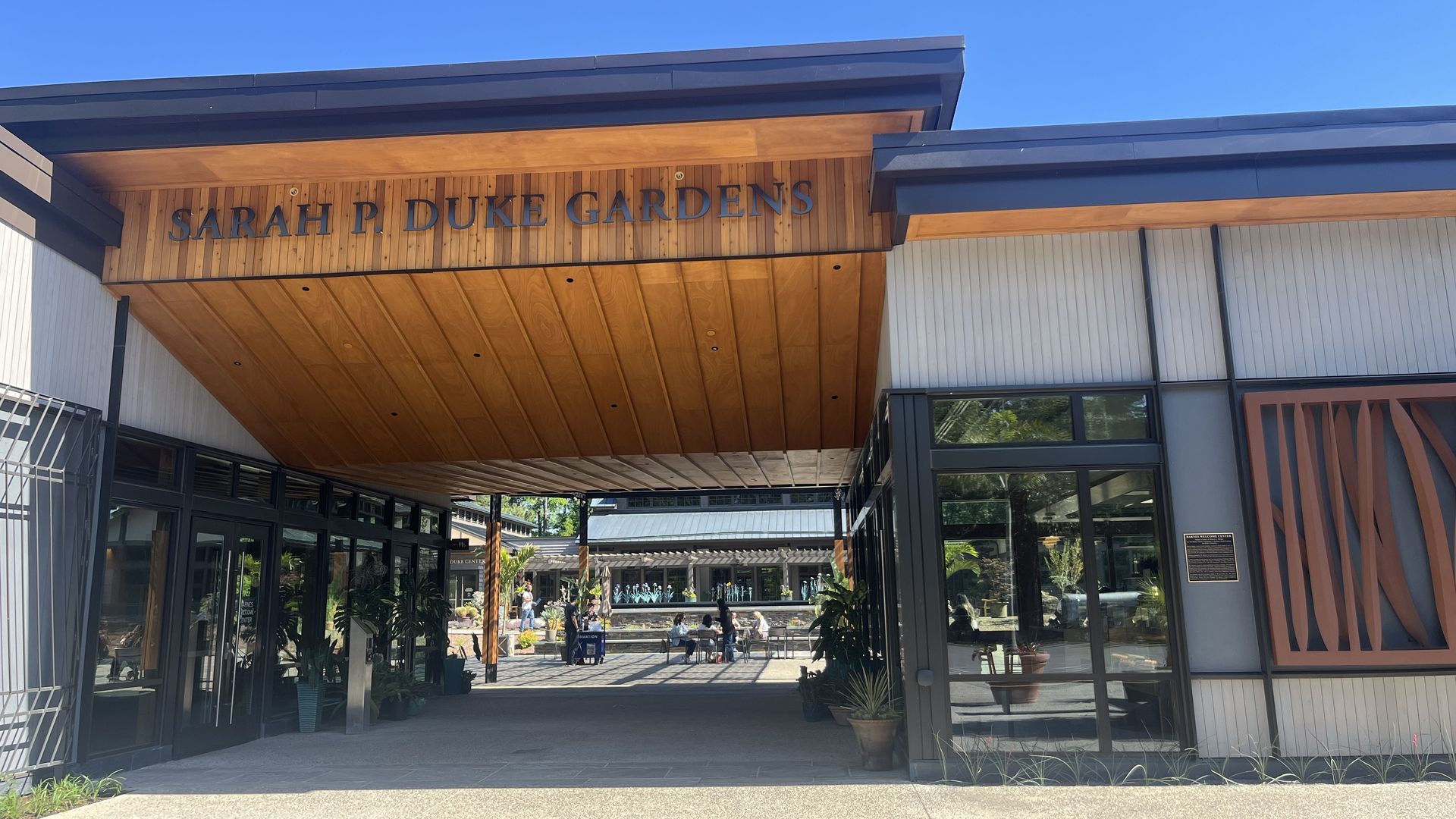 Entrance to "Sarah P. Duke Gardens" with a wooden overhang, glass doors, planters, and a bright blue sky.