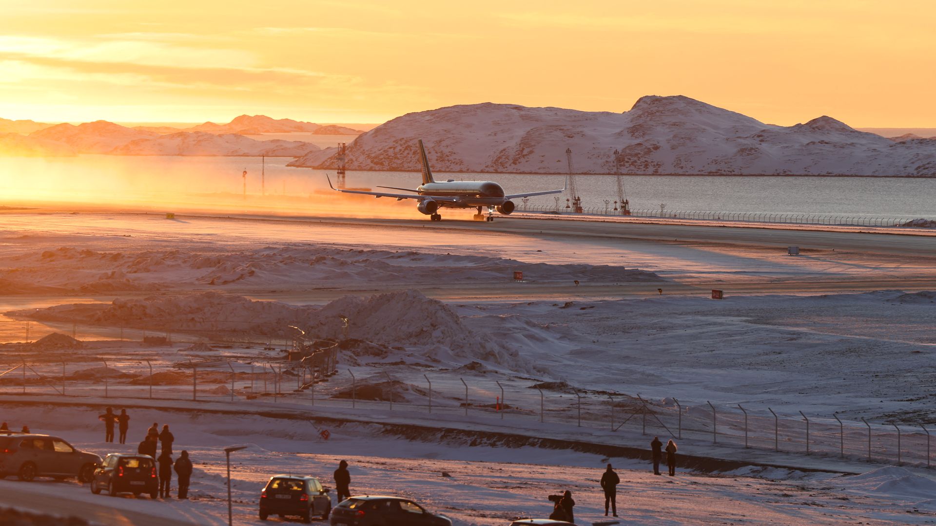 A plane lands on a runway in Greenland. People watch from afar, behind a fence. The sun is rising. It's snowy and icy.