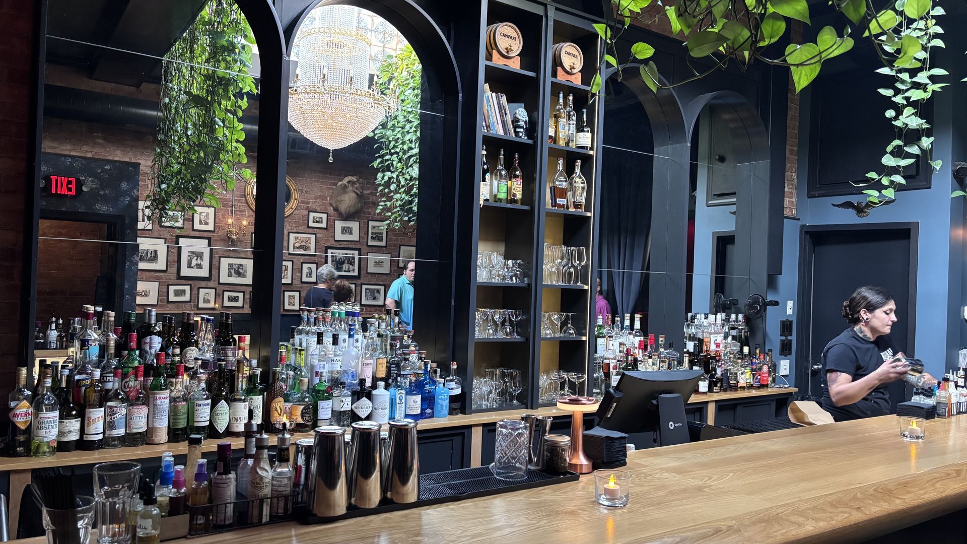 A chic bar interior with a long wooden counter full of bottled liqueurs, black metal arches, and hanging greenery. A crystal chandelier hangs above, brick walls with photos, and a bartender on the right.