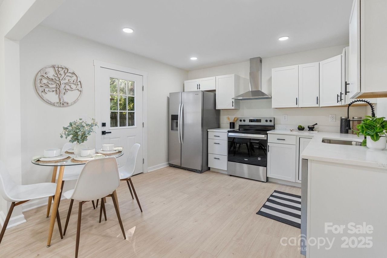 Bright modern kitchen with white cabinets, stainless steel appliances, light wood floor, small round glass dining table set for four, and a door with window. Decorative wall art and plants present.