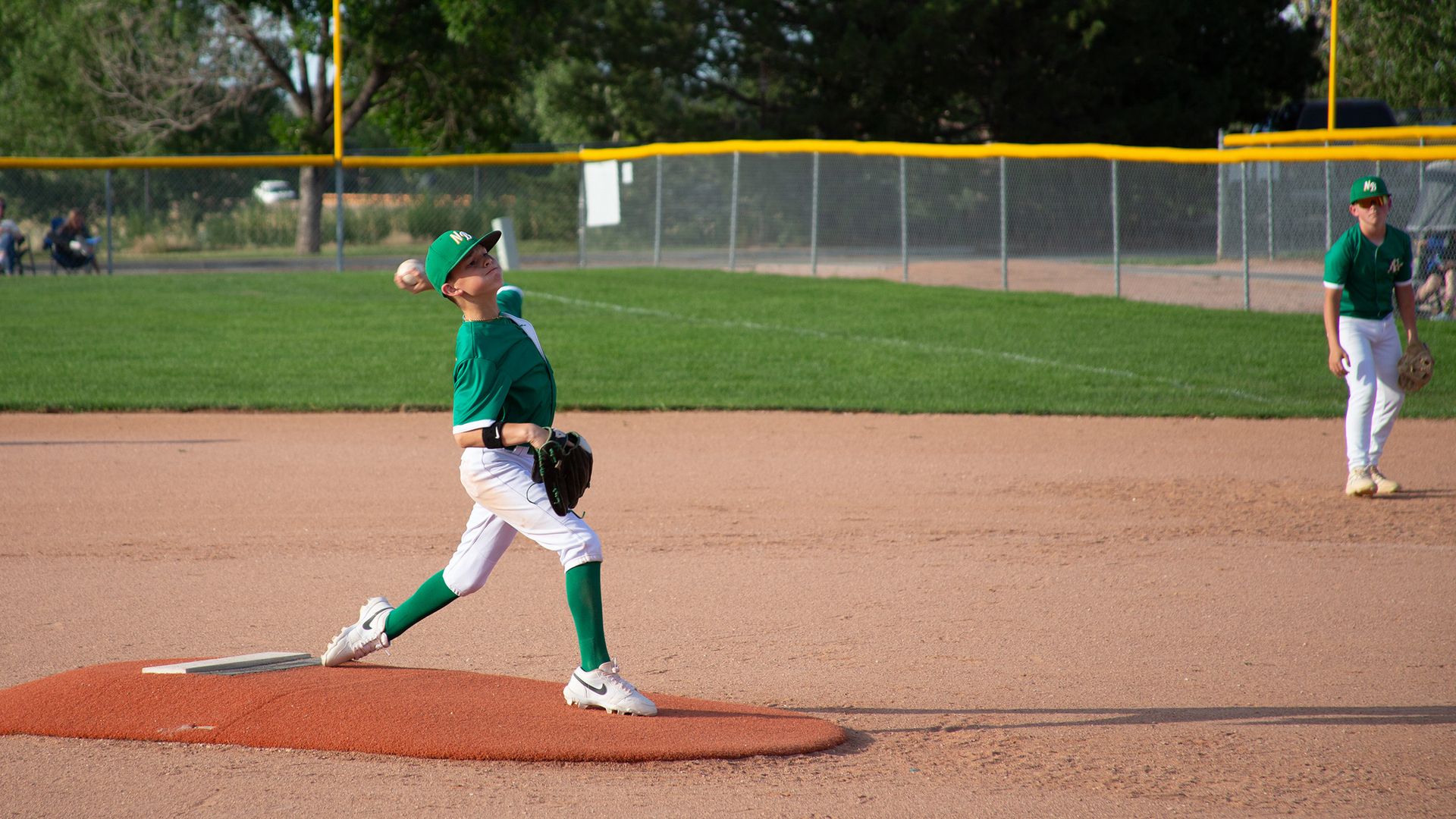 A youth baseball pitcher in a green jersey and cap throws a ball on the mound, with a teammate in green and white ready in the infield, and a fence and trees in the background.