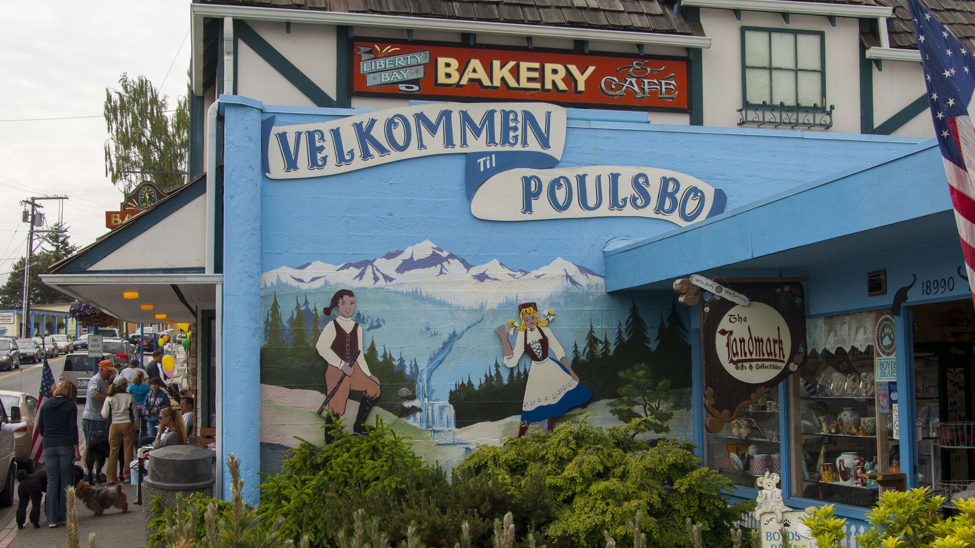 A street scene with a blue wall mural that says Velkommen Poulsbo with painted mountains, and people walking on the sidewalk nearby.