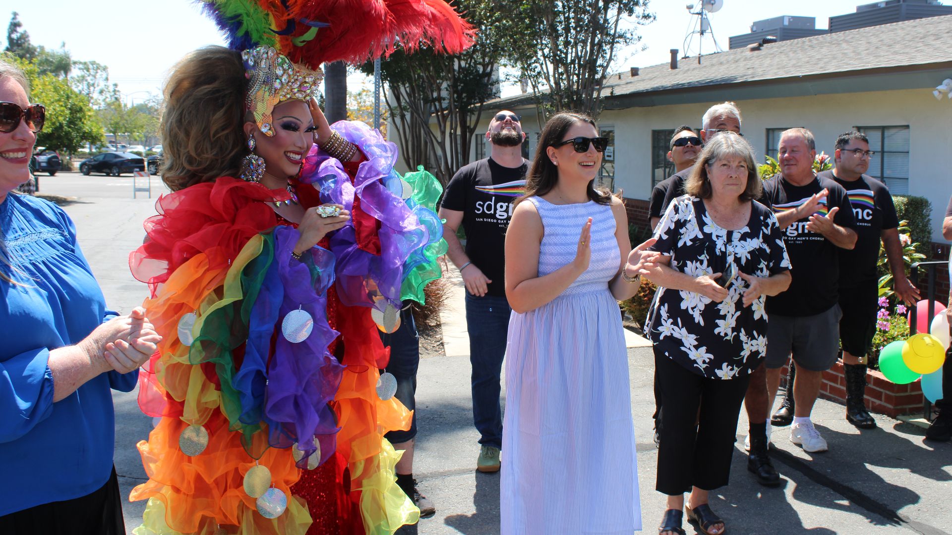 Colorful drag performer with an elaborate rainbow feather headdress and sparkling crown, wearing a red-and-rainbow ruffled gown, surrounded by smiling spectators outdoors on a sunny day, balloons visible.