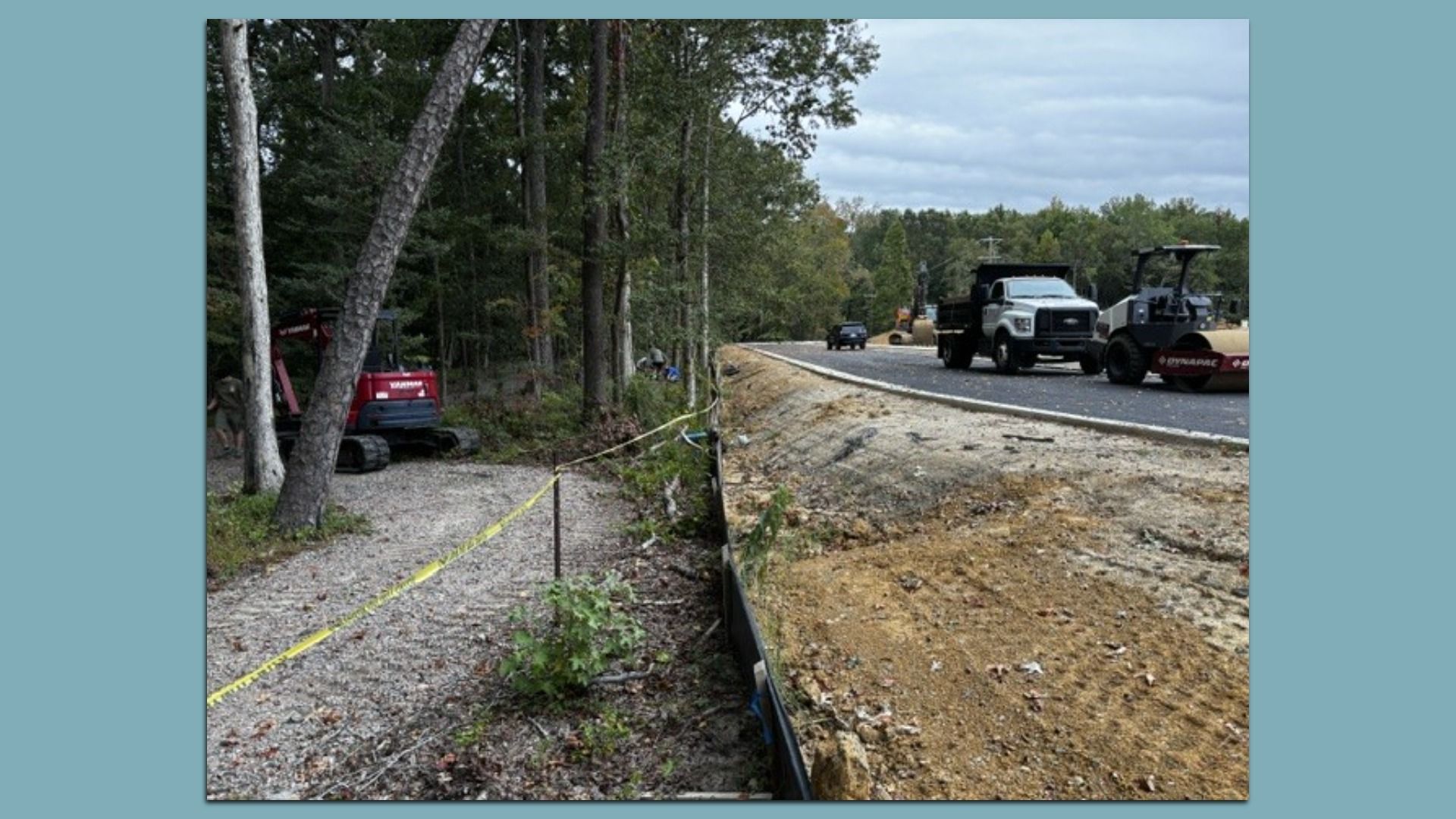 Road construction scene: left side trees with a red Yanmar excavator; right side a white dump truck and a road roller; yellow caution tape along a dirt trench under a cloudy sky.