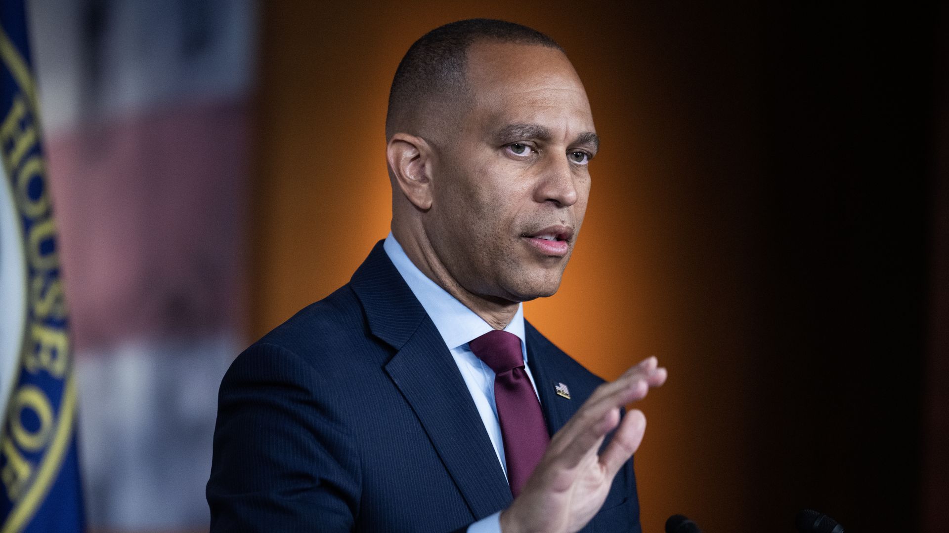 Hakeem Jeffries, wearing a blue suit and standing in front of an orange wall with his hand raised.