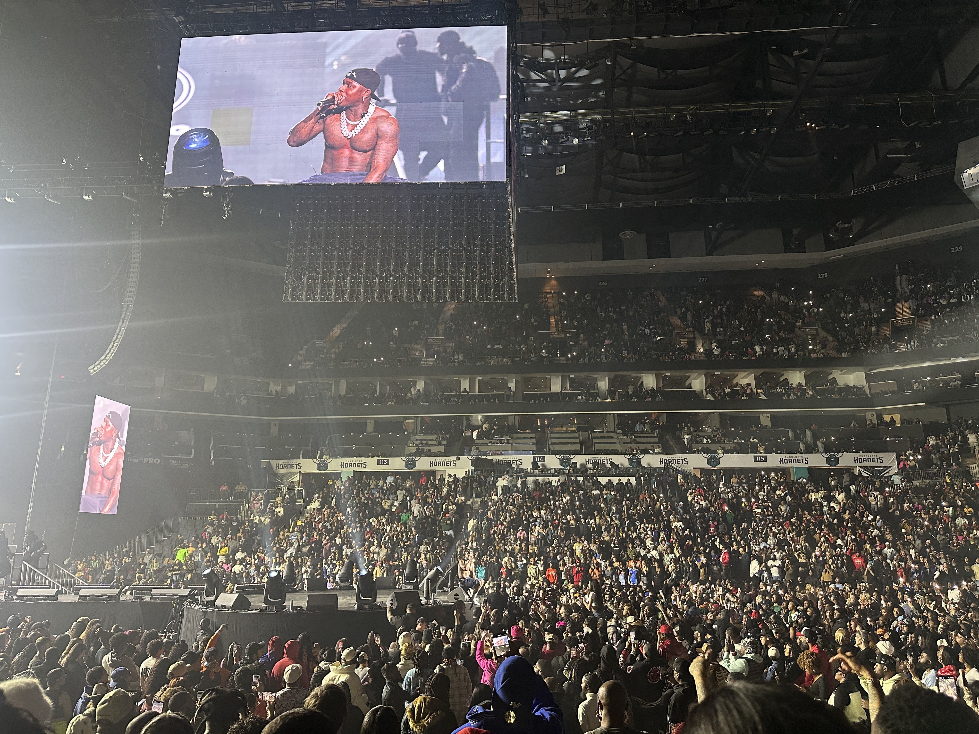 A packed indoor concert with a shirtless rapper wearing a chunky chain performing onstage; a large video screen above shows him as fans fill the arena.