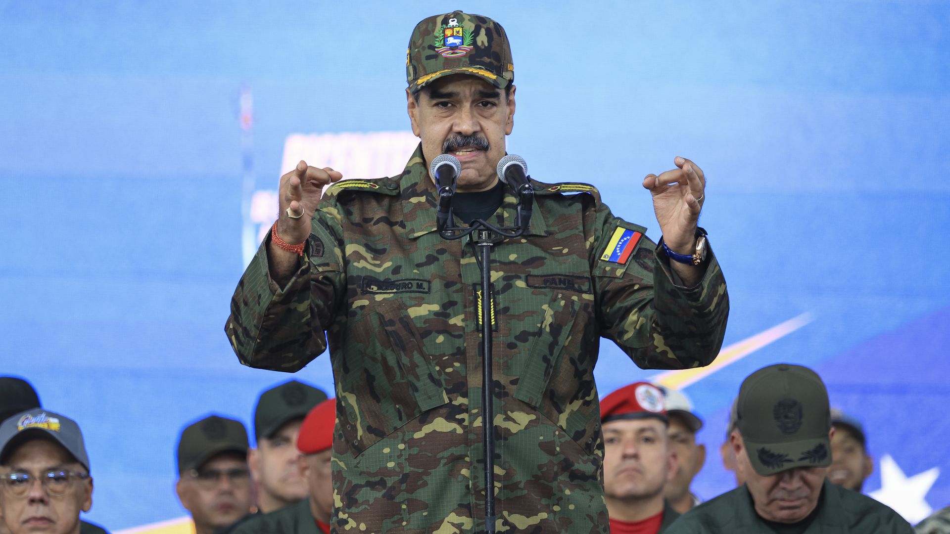 Nicolas Maduro, in military fatigues, gestures with both hands while a crowd sits behind him. 
