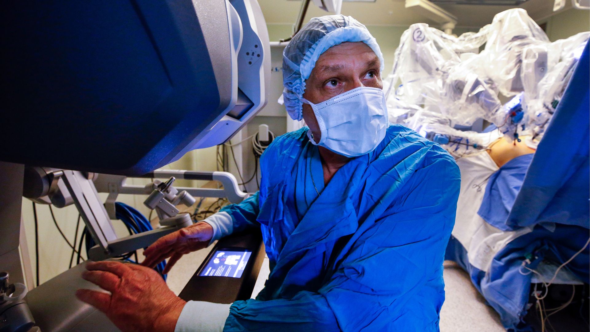 In this image, a surgeon in blue scrubs and wearing a white face mask looks to the right while using a machine during an operation.