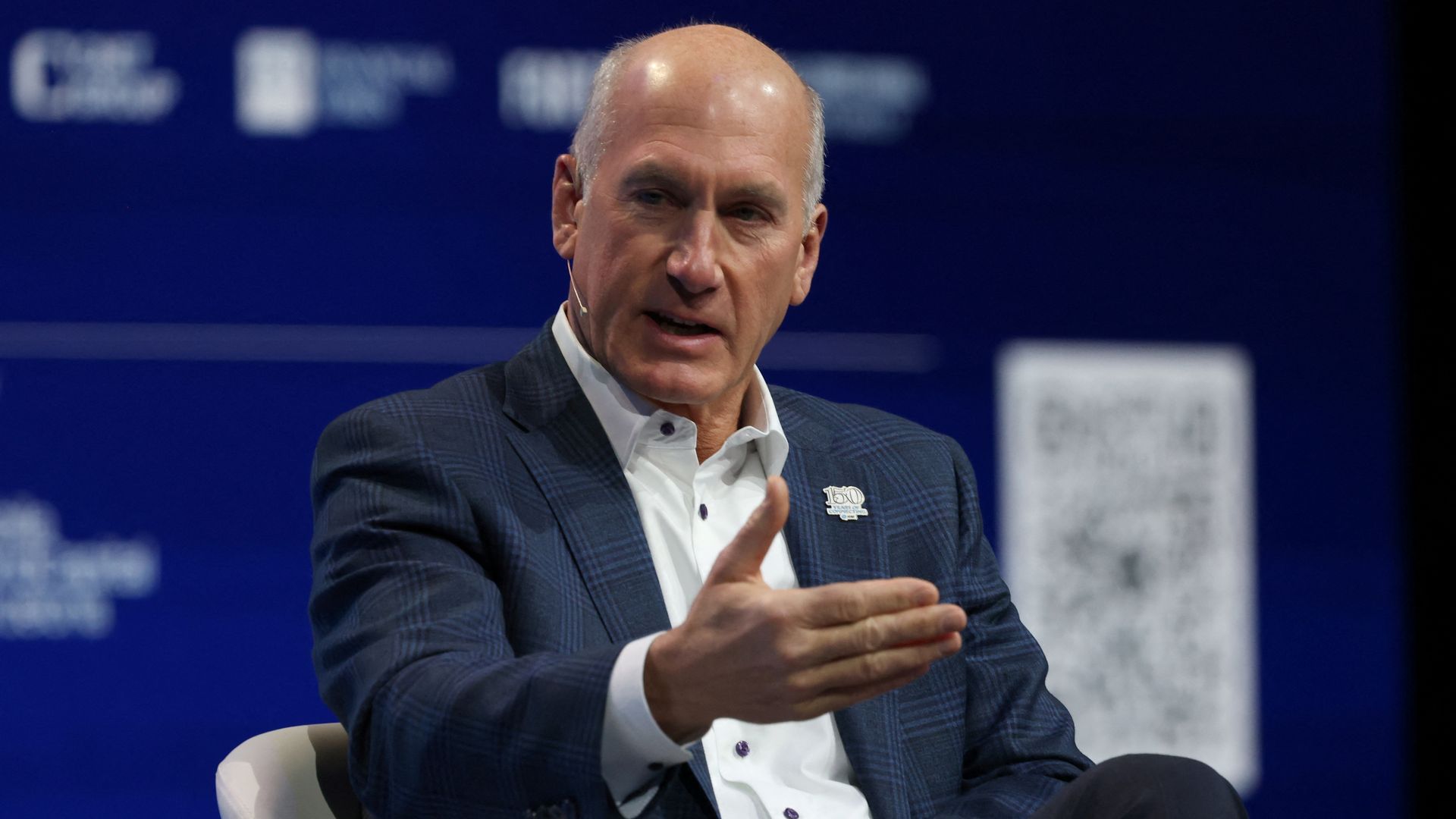 A person in a blue checkered blazer and white shirt sits on stage, wearing a headset microphone and gesturing with his hands as he speaks against a blue backdrop.