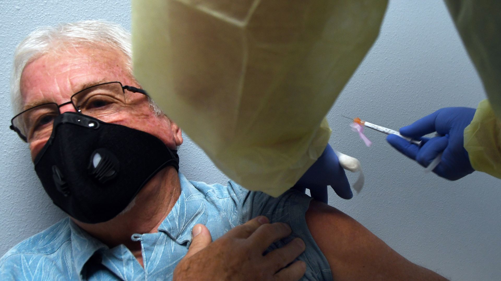 A vaccine syringe is injected into the arm of a man who is wearing a mask.