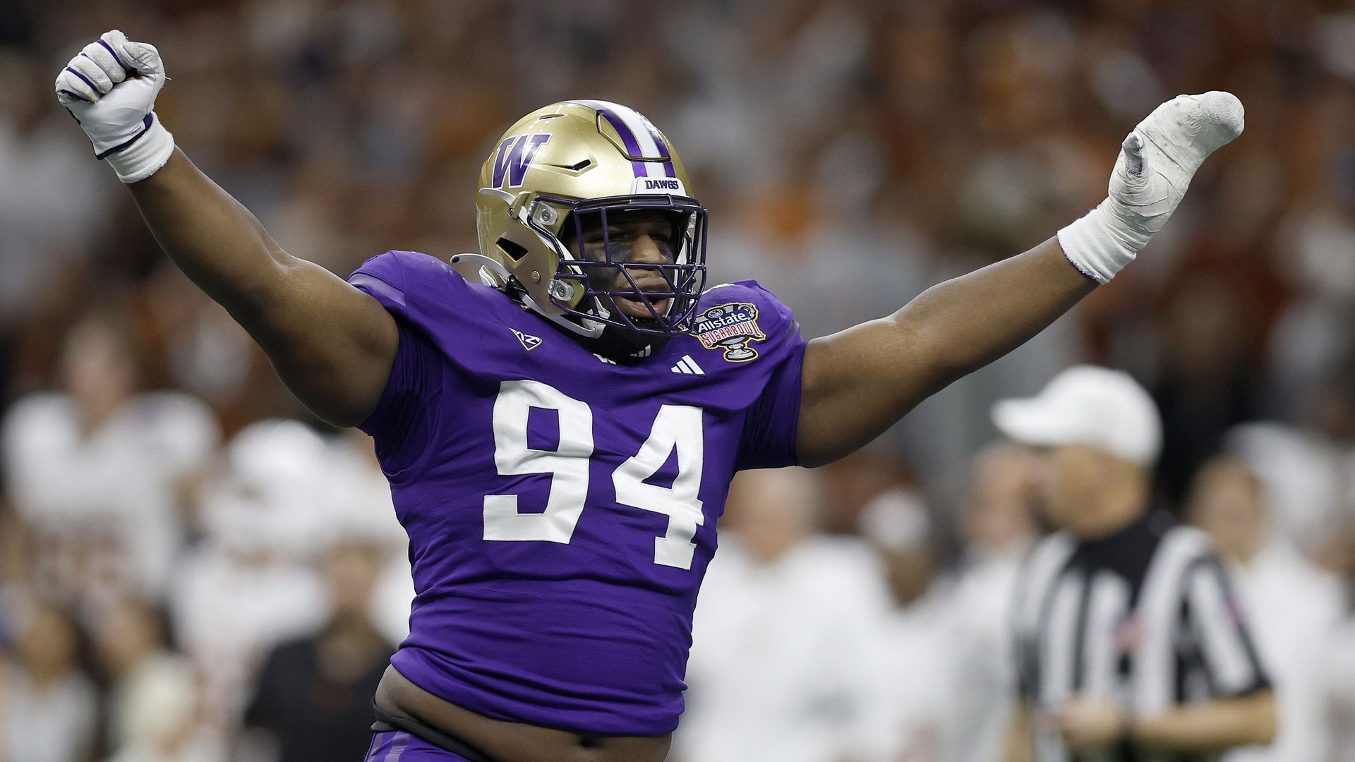 A player in a purple jersey and gold helmet with the number 94 on his jersey holds up both hands in a V formation over his head, with a stadium crowd blurred in background.