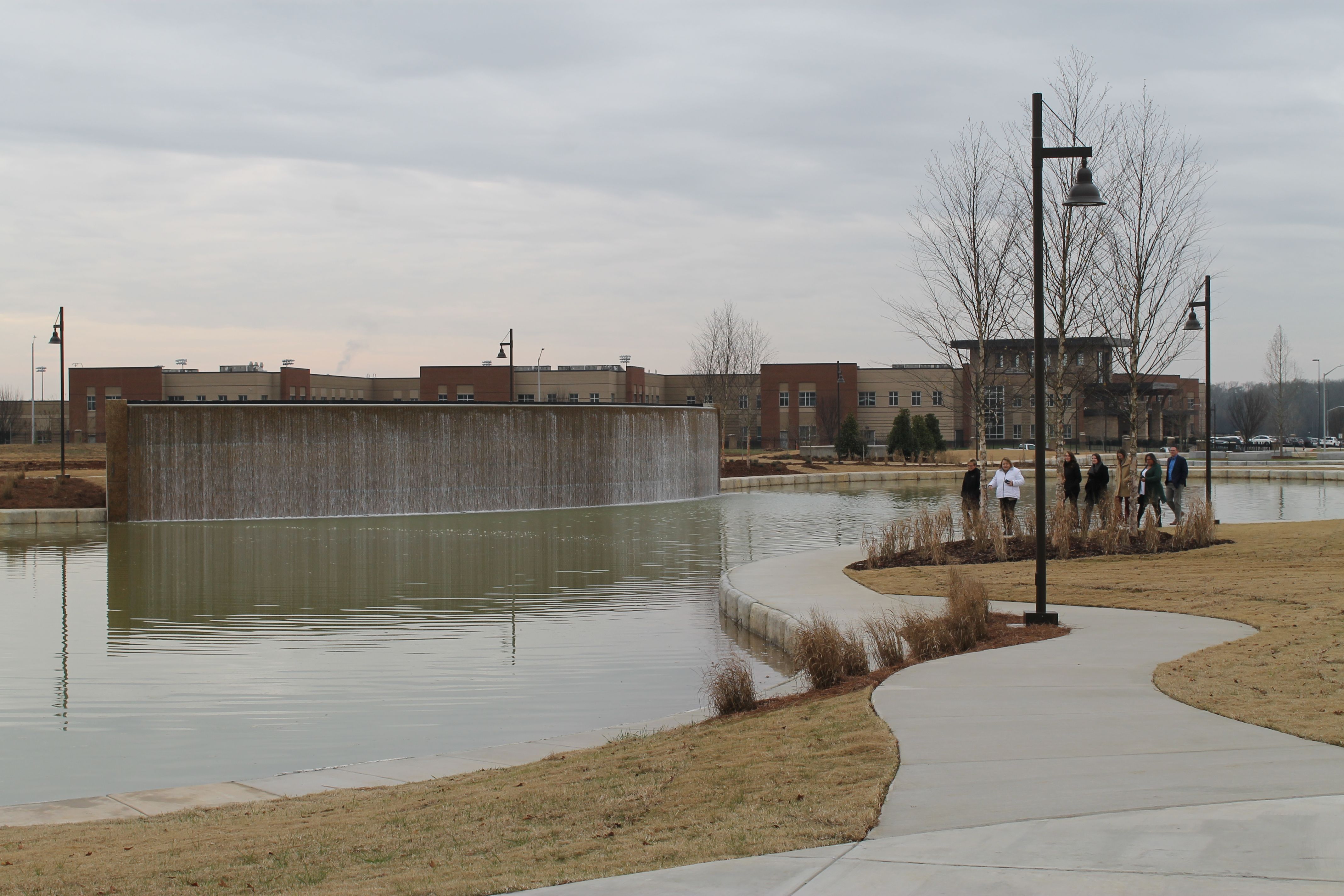 People walking on a curved sidewalk beside a pond with a large rectangular waterfall and a beige and red brick building in the background under an overcast sky.
