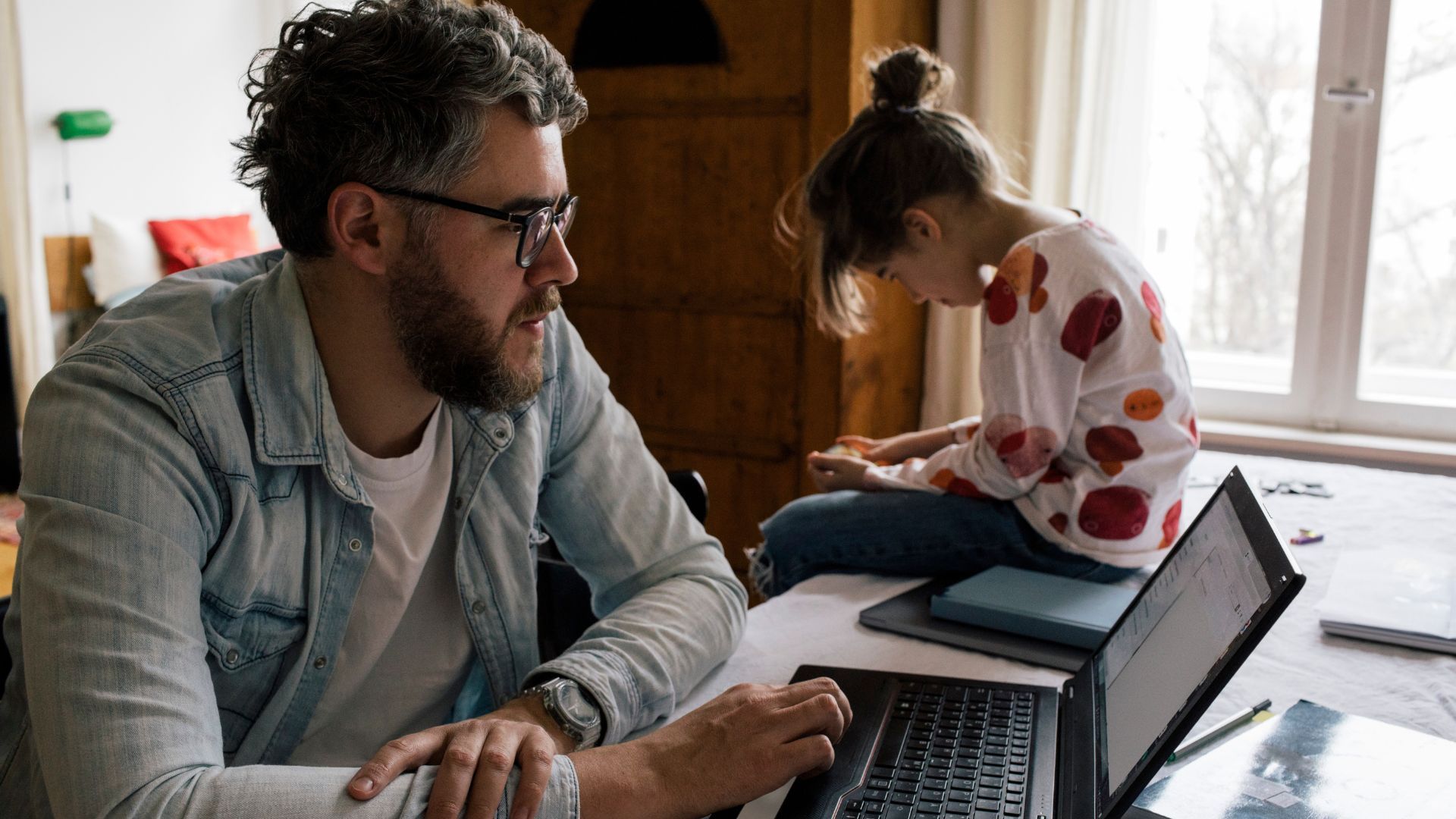 A dad looking at his laptop with a child sitting next to him.