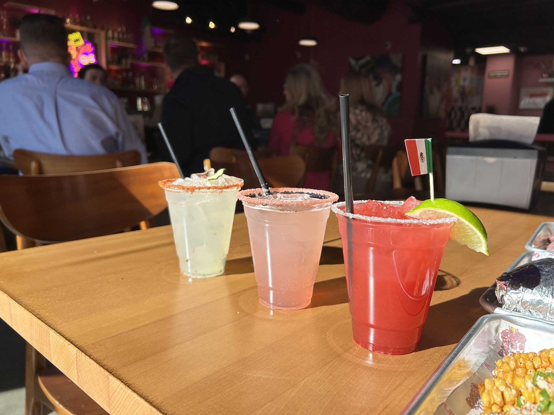 Three colorful cocktails on a wooden table: a pale green drink, a pink drink, and a red drink with lime, plus a tiny Mexican flag; blurred bar and customers in the background.