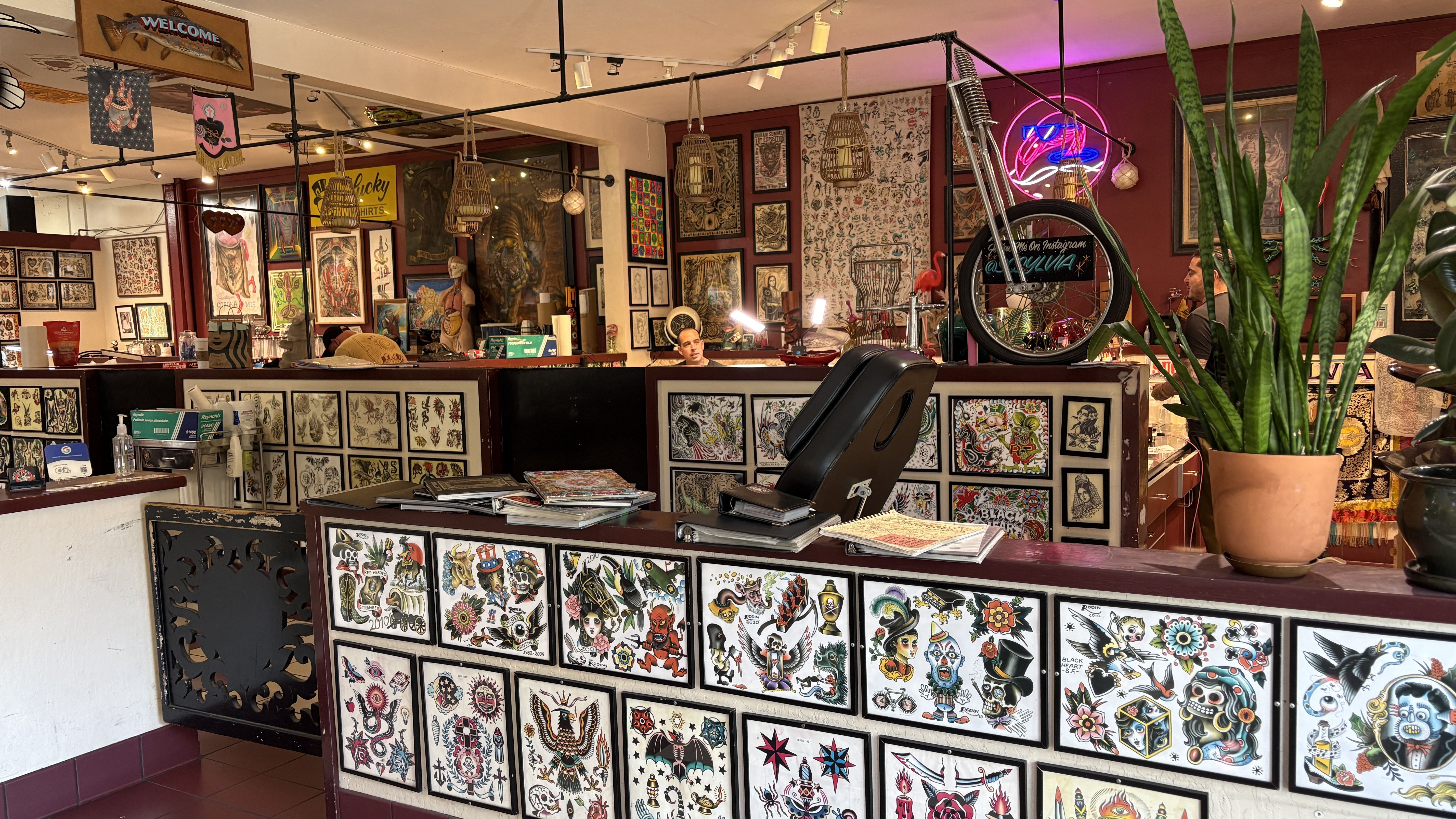Colorful tattoo studio filled with framed flash art on walls and front counter; hanging lanterns, a neon sign, a bicycle, and a large potted plant on the right as artists work in the back.