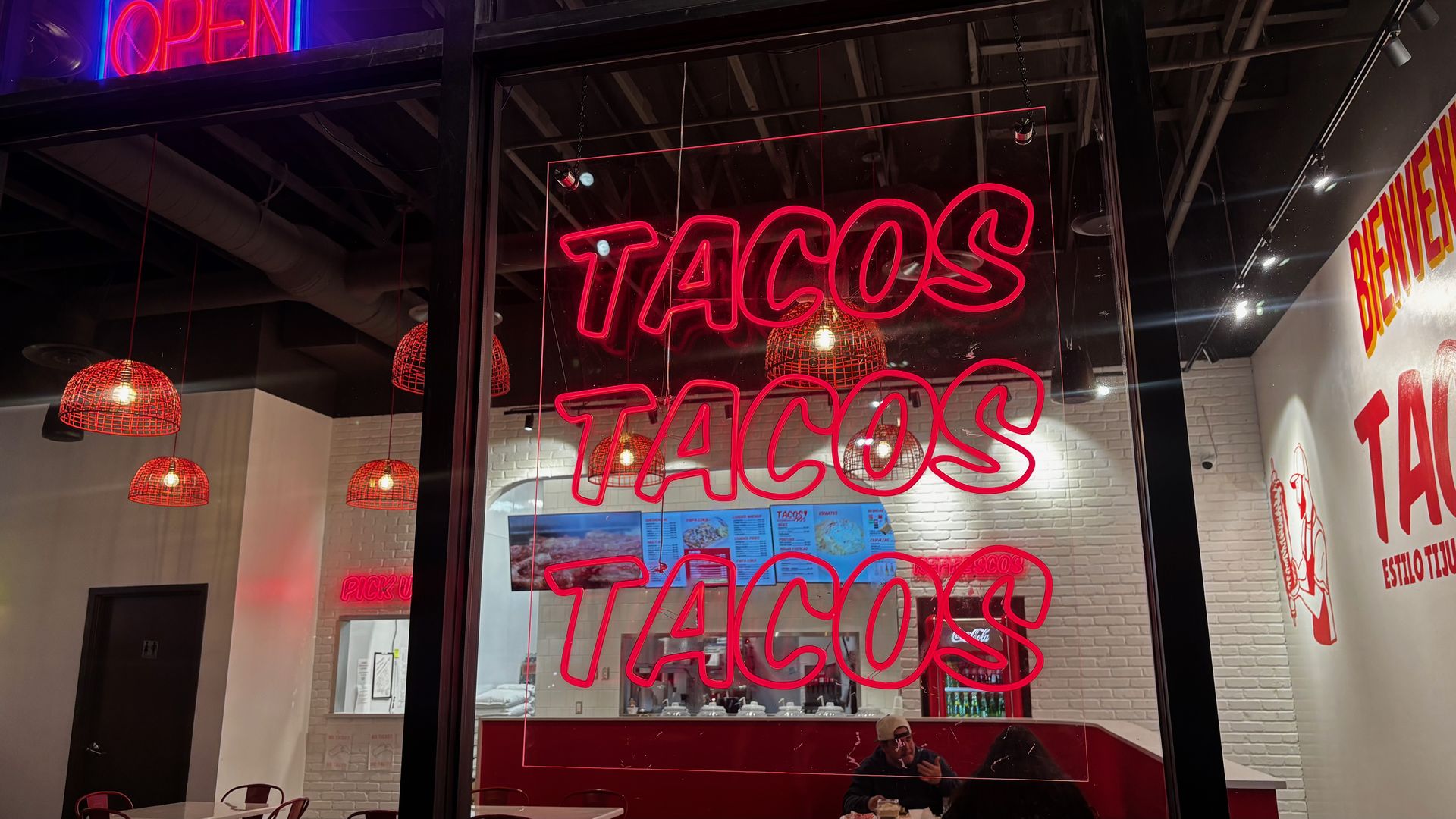 Interior of a taco restaurant at night with red neon signs reading "TACOS TACOS TACOS" and "OPEN," red hanging lights, a menu on the wall, and customers sitting at tables.