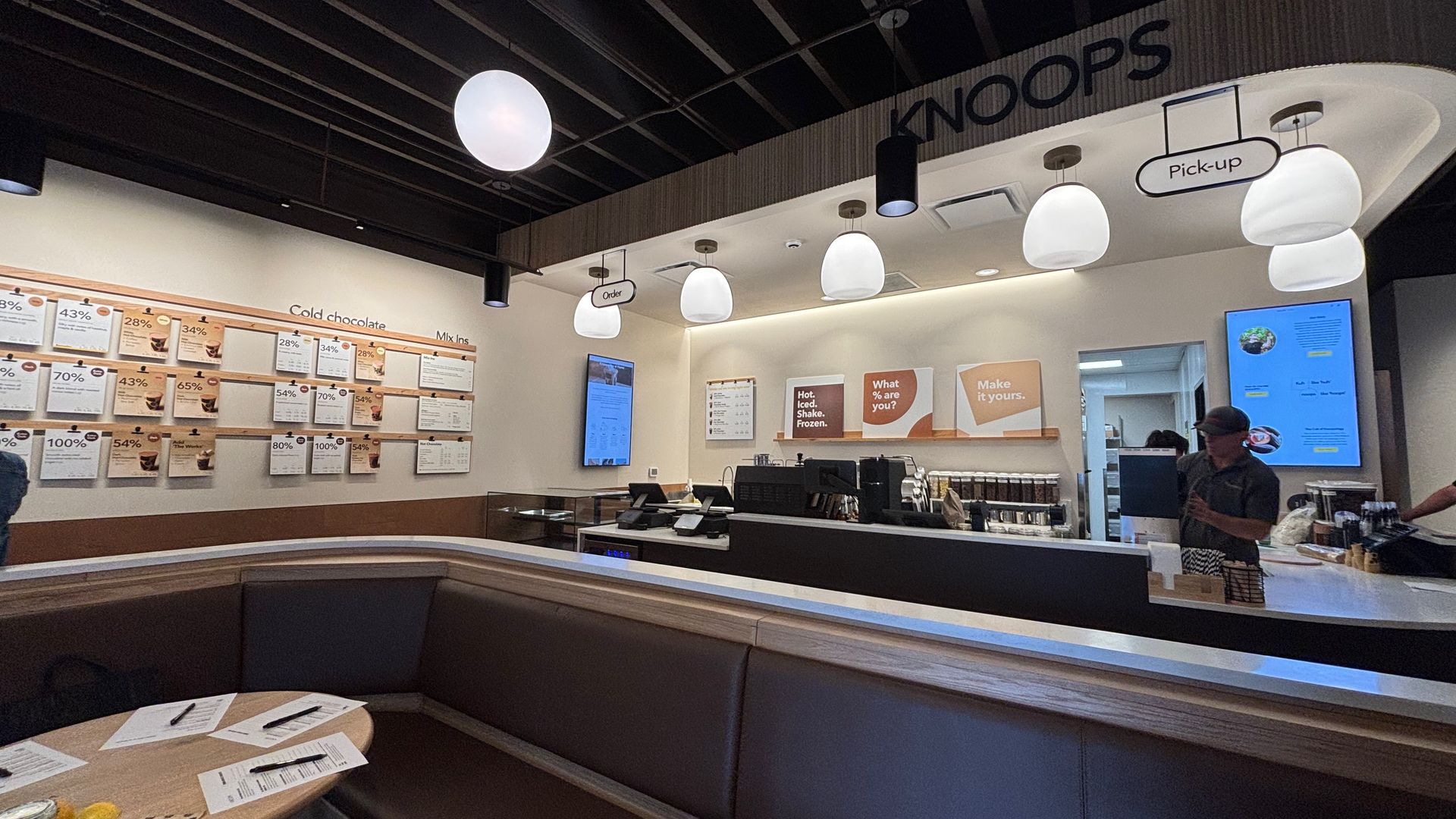 Cafe interior with a black ceiling and round white pendant lights, curved brown seating along a long counter, wall menu boards, and signs reading KNOOPS and Pick-up above the counter.
