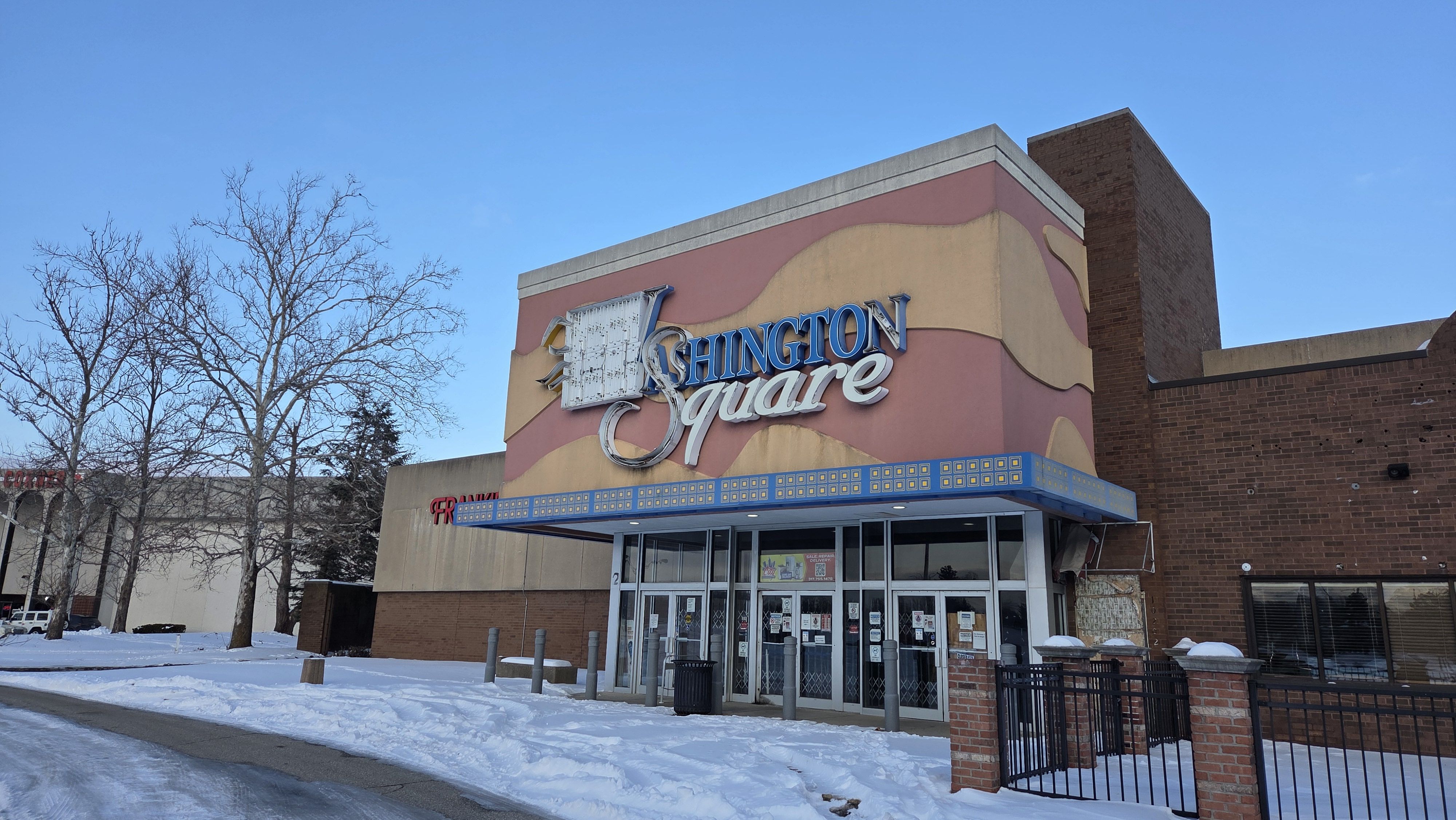 Clear blue sky over a retro storefront with pink and tan curved facade, reading "Shington Square" in blue script. A blue and yellow marquee, leafless trees, and snow on the ground.