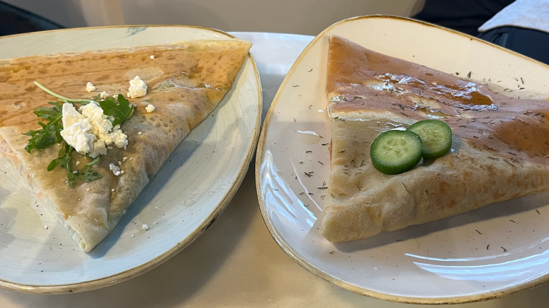 Two savory crepes, one with cucumber slices and the other with cheese, sit on  plates at a table. 