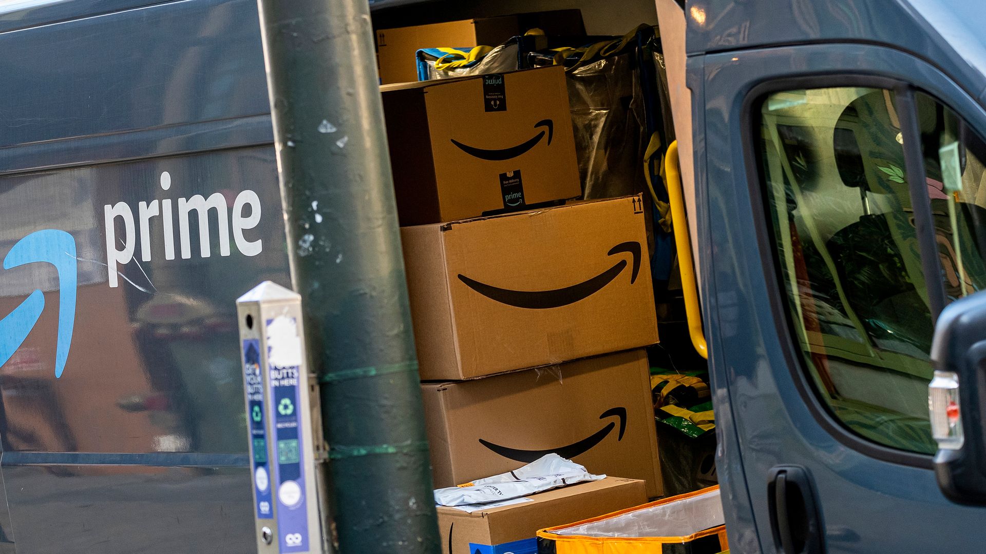 Open Amazon Prime delivery van with stacked Amazon boxes and a yellow carrier bag inside, parked next to a pole with cigarette butt disposal unit.