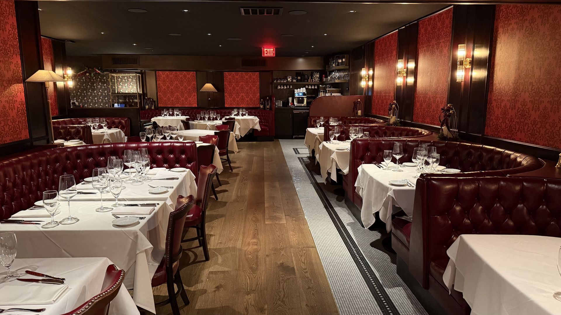 Restaurant dining room with red leather booths, white tablecloths, set tables with glassware and silverware, warm lighting, red patterned wallpaper, and wooden flooring.