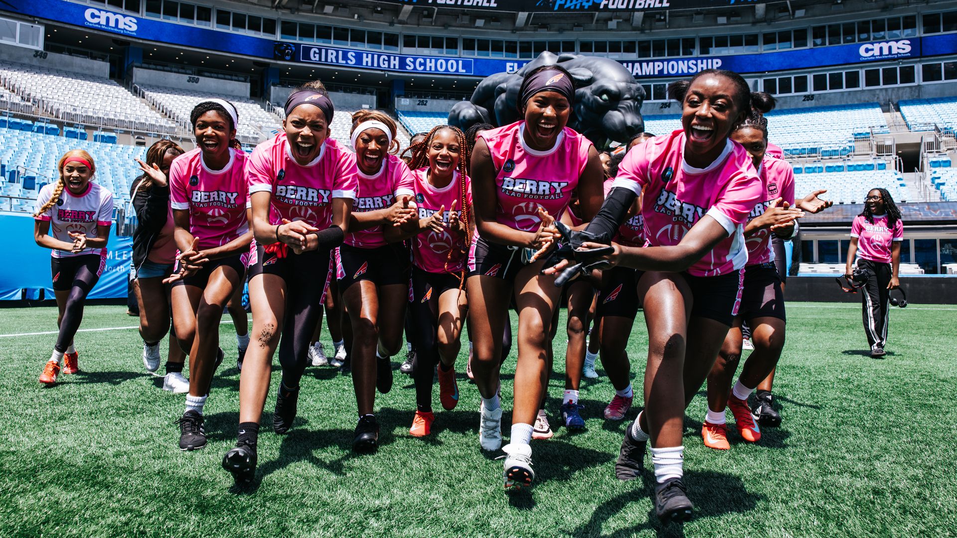 Group of girls in pink flag football uniforms sprint toward the camera on a green turf field inside a large stadium with blue seating and banners reading "Girls High School Championship".