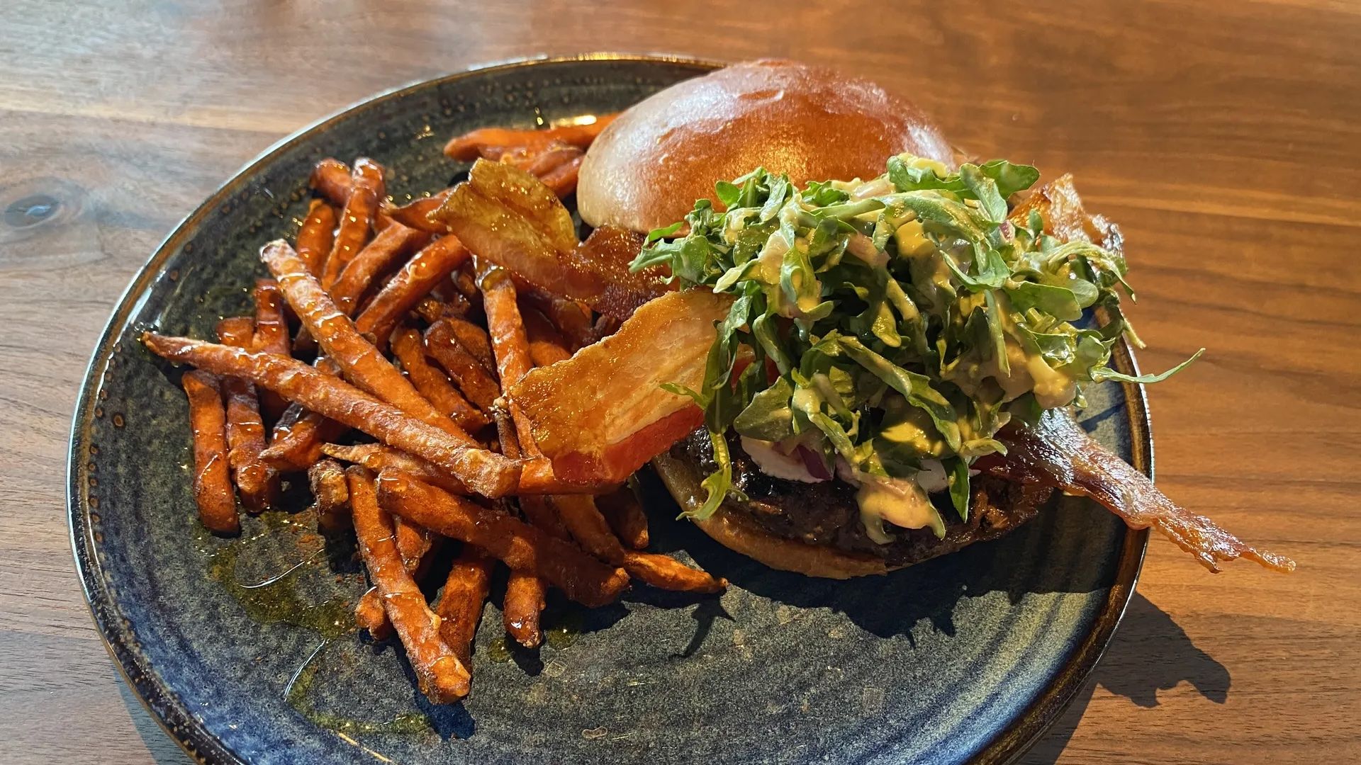 Plate with a bacon-topped burger with greens and sauce, served alongside a pile of crispy sweet potato fries on a dark ceramic plate over a wooden table.