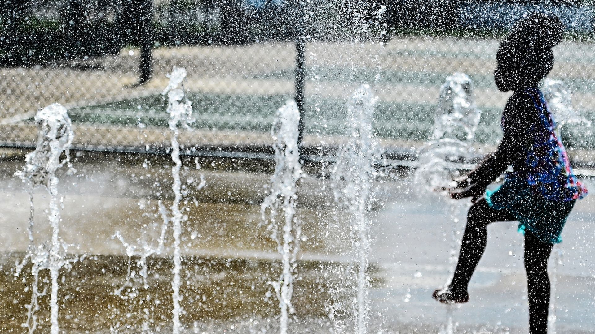 Silhouette of a child in a colorful outfit playing and kicking water jets from a splash pad on a sunny day with a chain-link fence in the background.