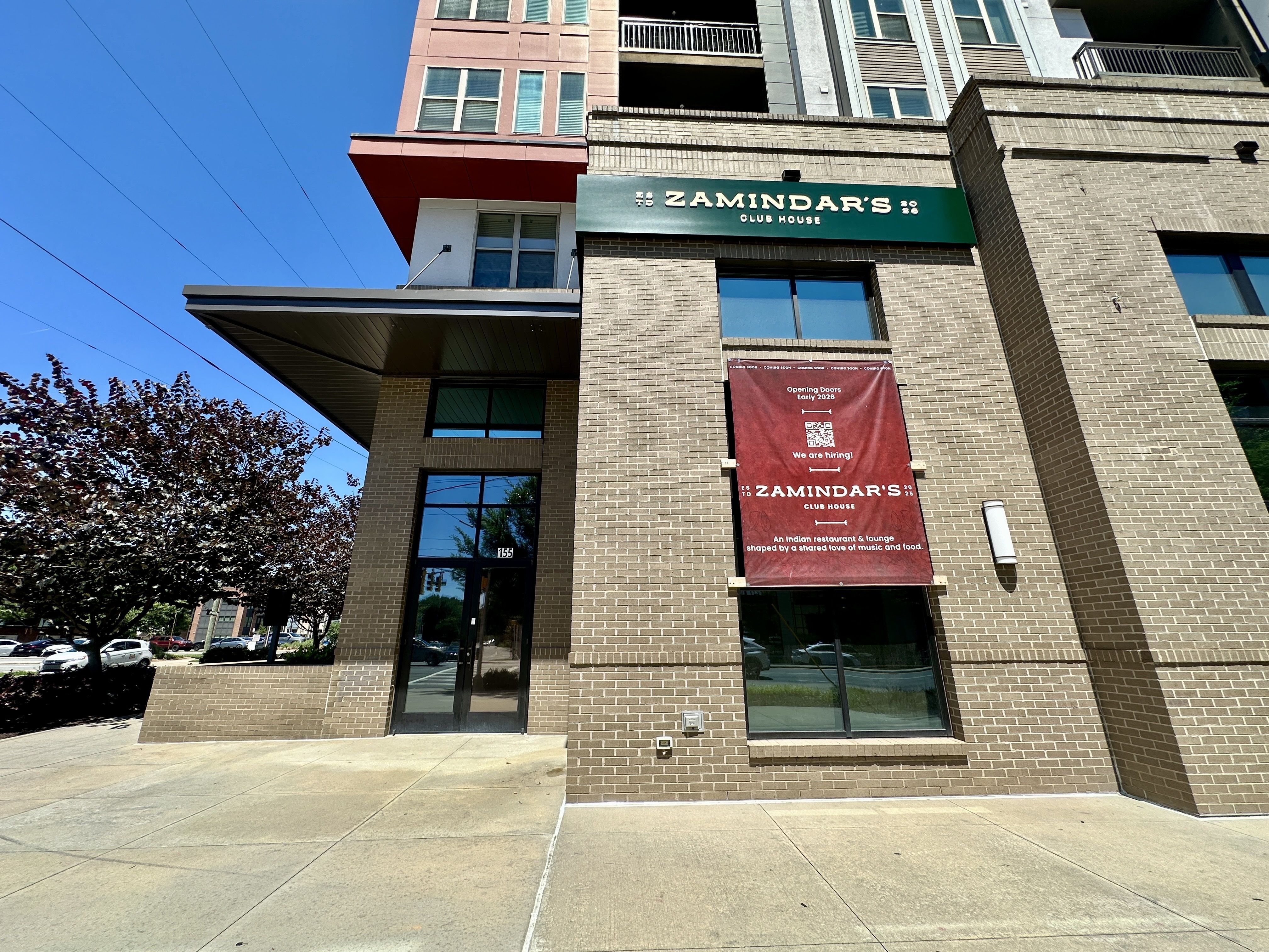 Brick building with a green sign reading "THE ZAMINDAR'S CLUB HOUSE" above glass doors; red banners, blue sky, power lines, and a tree on the left.