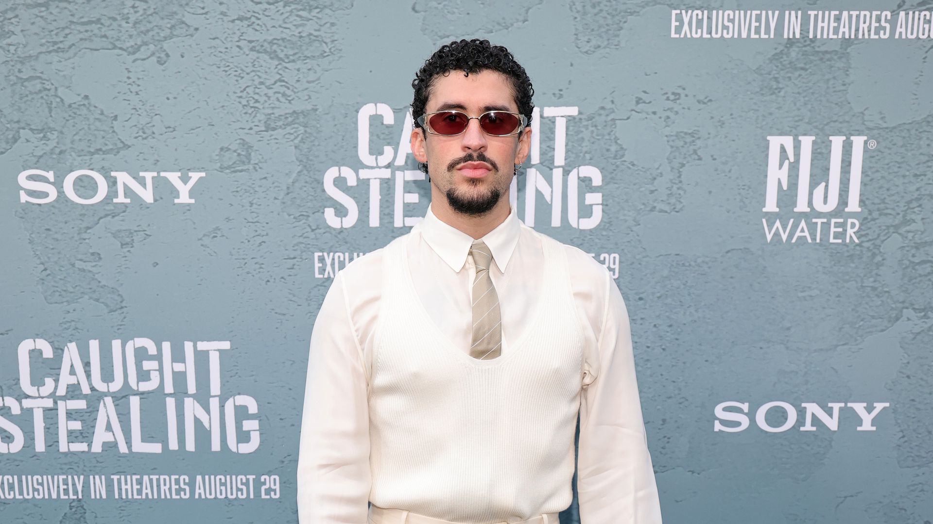 Puerto Rican singer Bad Bunny poses for a photo, wearing a fully white outfit with a beige tie. 