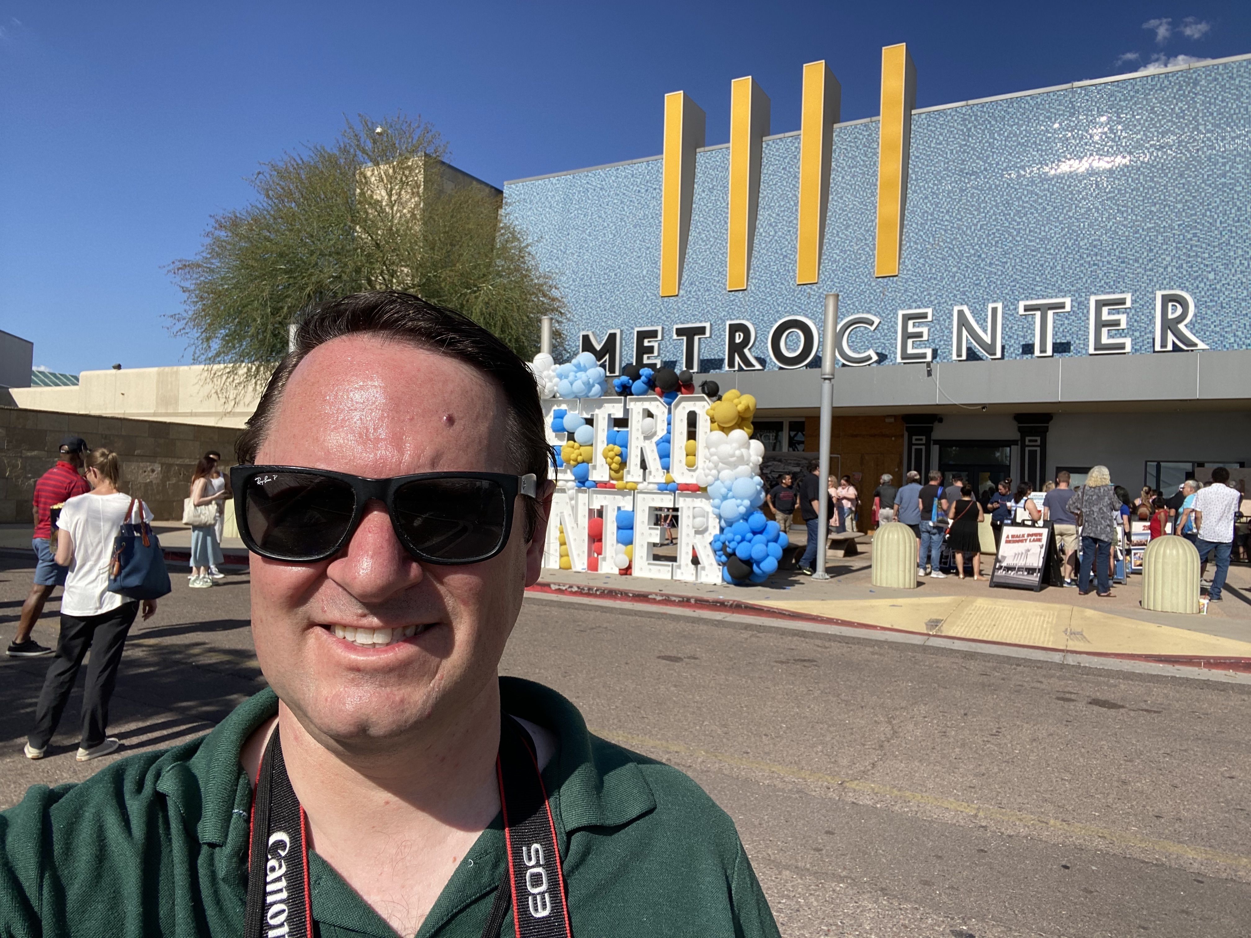 A man takes a selfie in front of a mall with a sign behind him that says Metrocenter. 