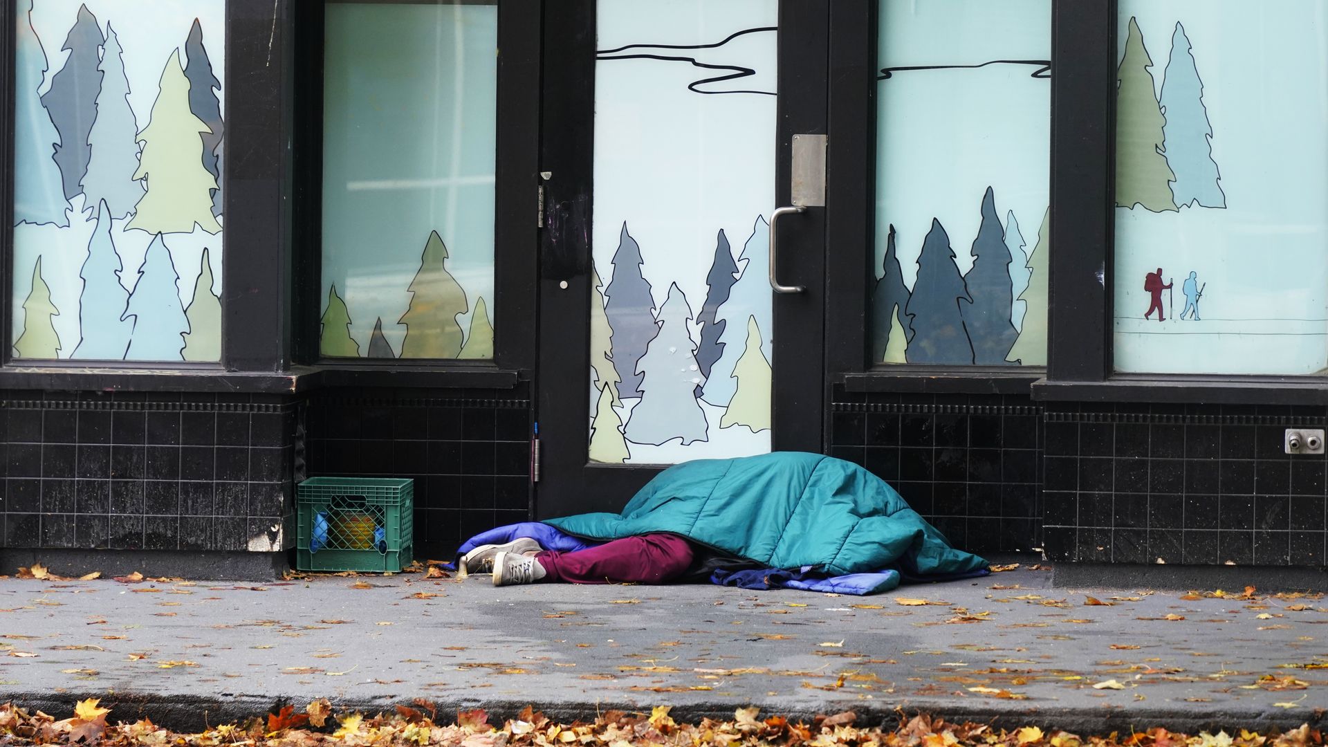 Storefront with black-framed glass doors and winter forest art on the windows; a person sleeps under a blue sleeping bag on the sidewalk, next to a crate, shoes, and scattered leaves.