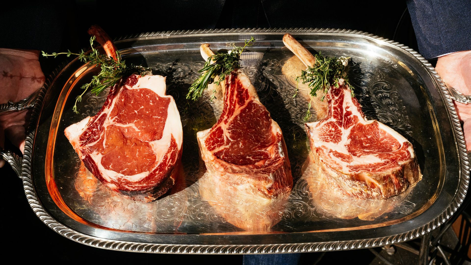 A trio of steaks sit on a silver platter that someone holds.