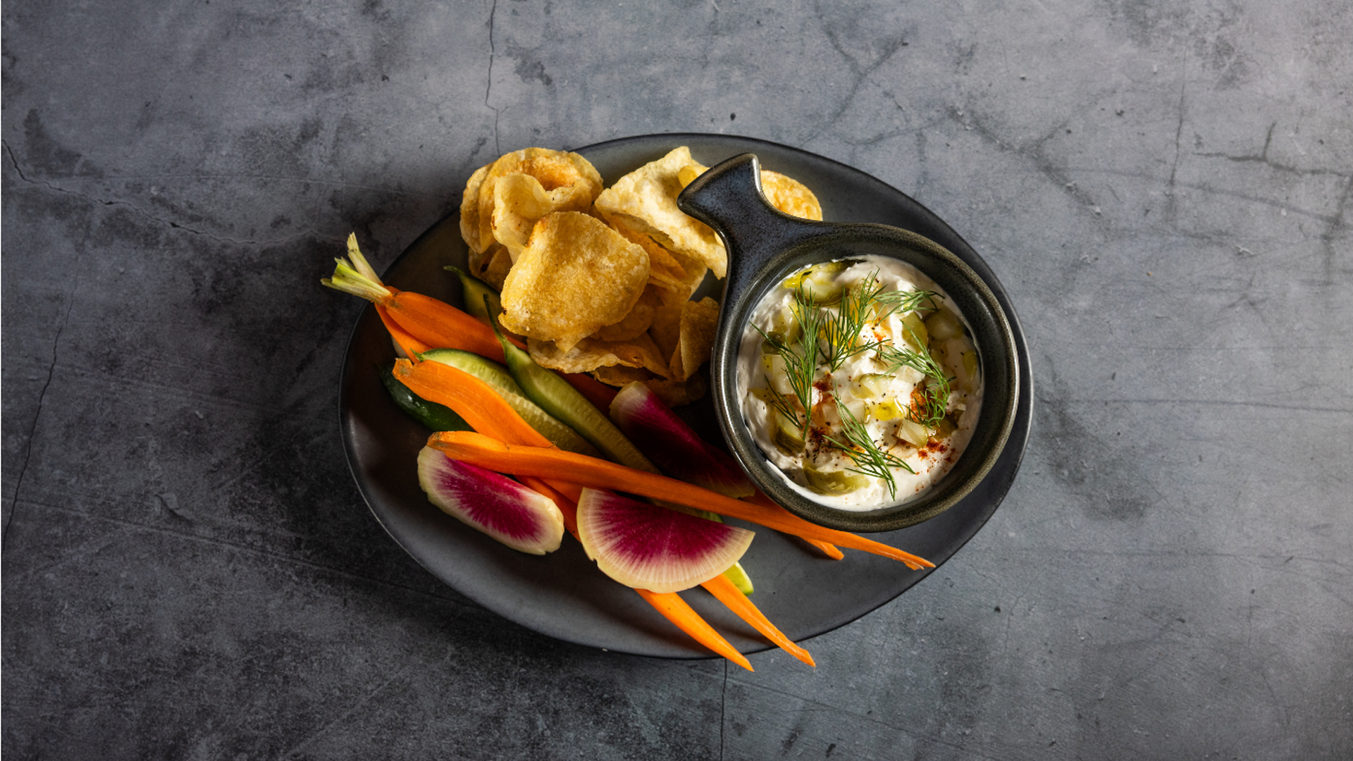 Top-down view of a dark plate with potato chips, carrot sticks, radish slices, cucumber, and a small pot of creamy dill-dusted dip on a gray stone surface.