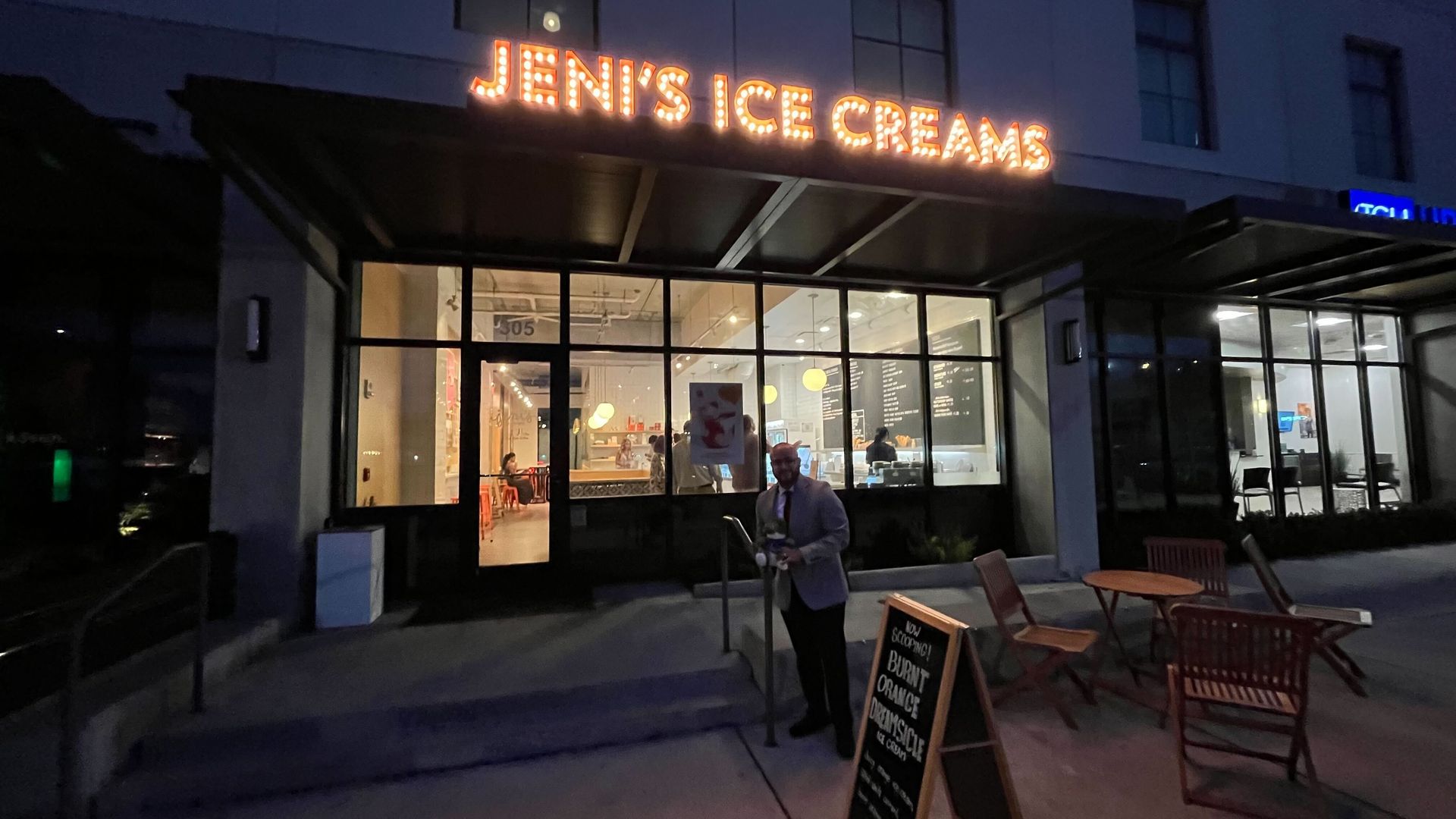 Nighttime storefront with a glowing marquee that reads "JEN'S ICE CREAMS". Glass windows reveal a warmly lit interior with customers; outdoor tables and chairs on the right, and a suited man at the entrance.