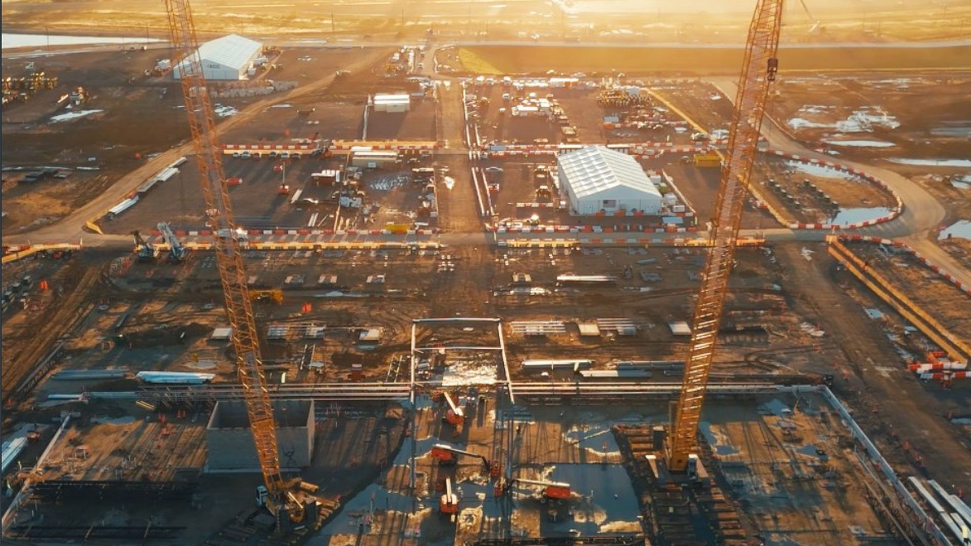 Aerial view of a large construction site at sunset: two tall yellow cranes dominate the foreground, white temporary buildings, barriers, and organized work zones spread across muddy ground.