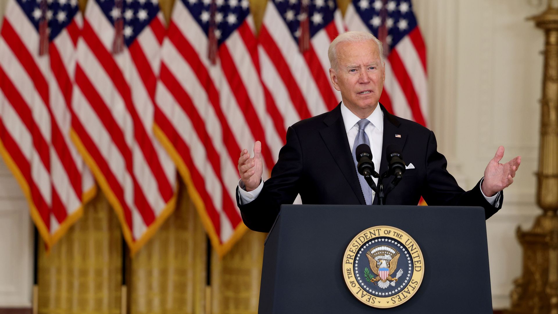 President Joe Biden stands in front of a row of American flags and gestures at a podium with an insignia reading President of the United States