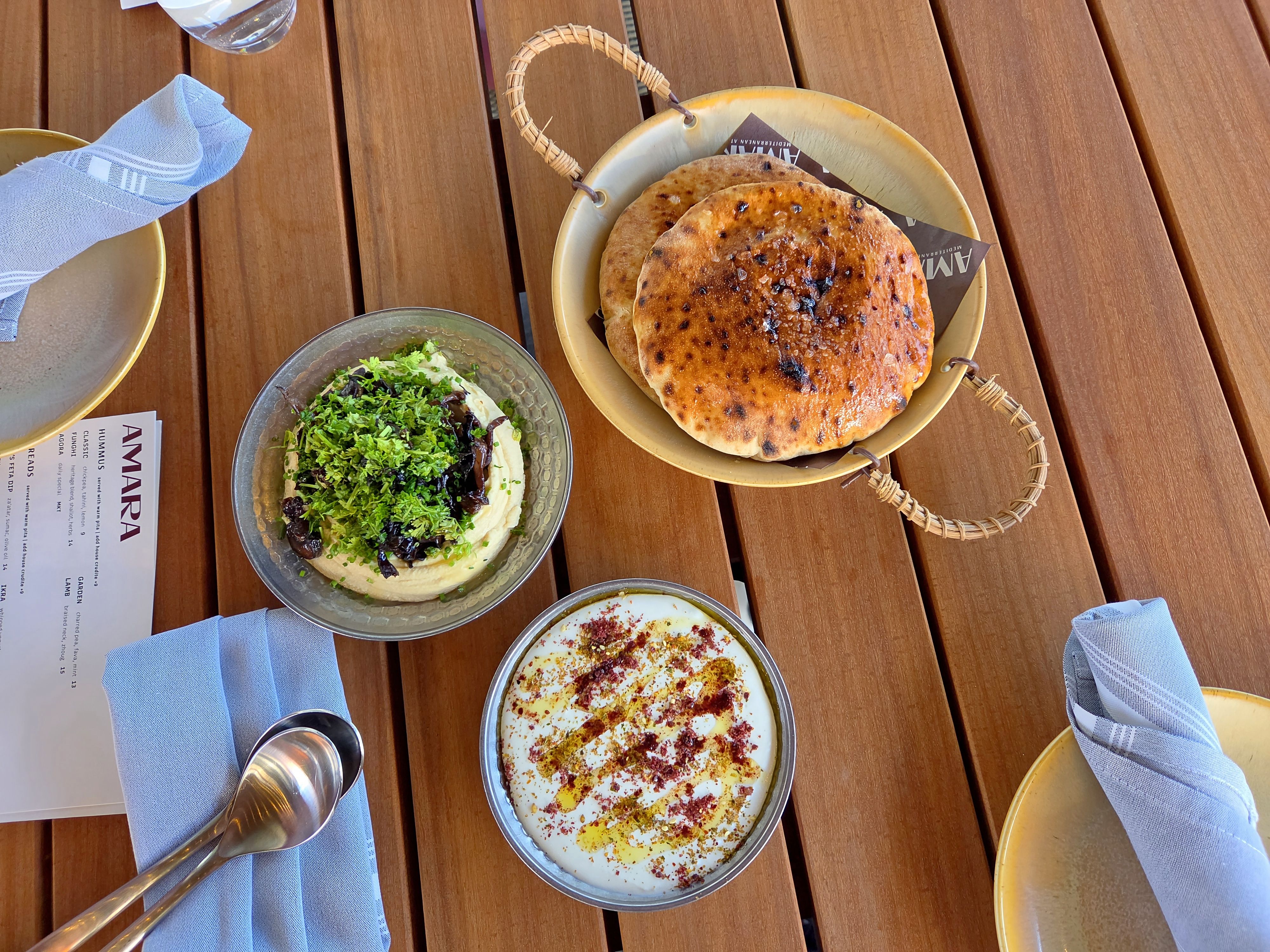 Top-down view of a wooden table with a wicker-handled basket of golden-brown flatbreads and a bowl of herb-topped hummus; blue napkins and other plates surround the meal.