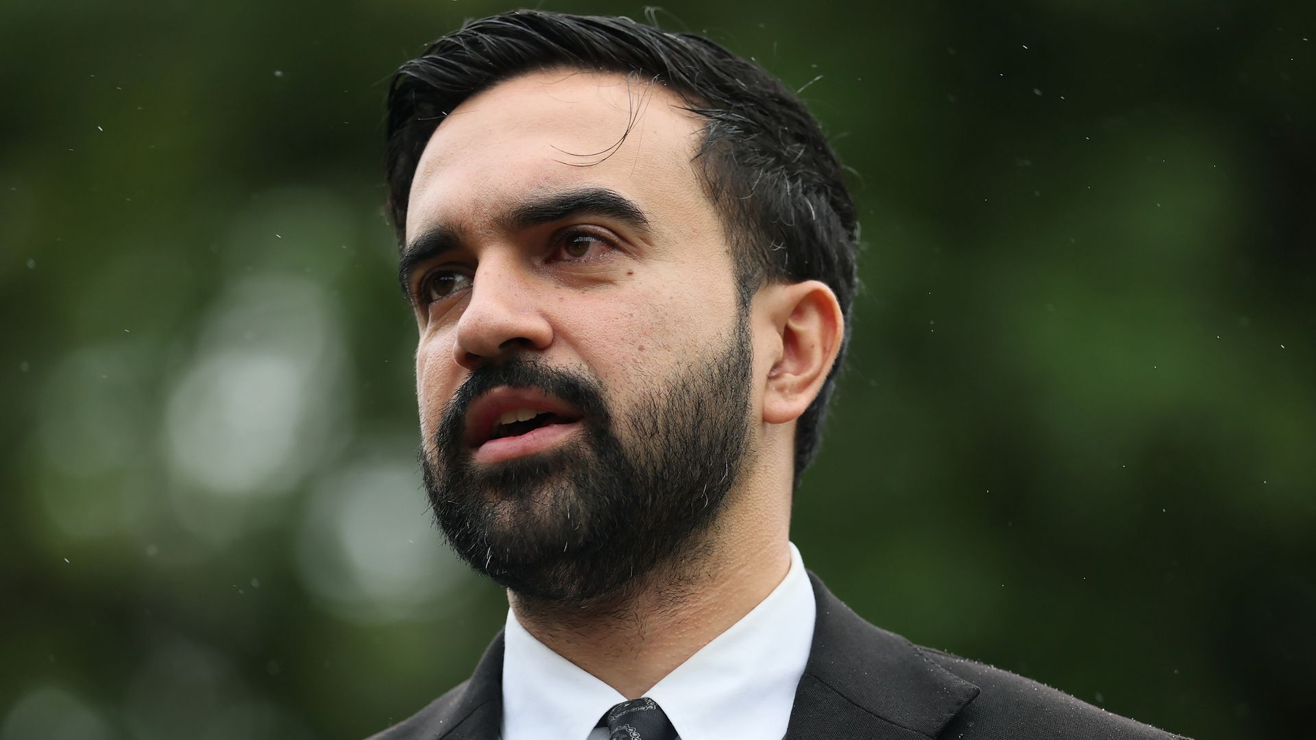 A black-haired and bearded New York Mayoral Candidate Zohran Mamdani speaking in St. James Park, NYC, wearing a black jacket, white shirt and black tie.