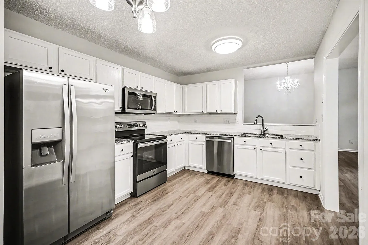 Bright L-shaped kitchen with white cabinets, granite countertops, and stainless steel appliances. Wood-look flooring, a stainless sink, and a chandelier in the adjacent dining area.