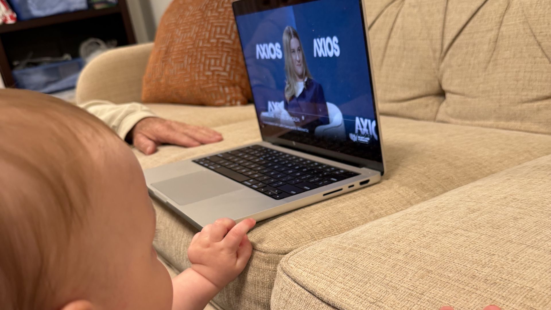 Close-up of a small child in a blue shirt reaching toward a silver laptop on a beige tufted sofa. The screen shows a woman presenter on a blue background with white text.