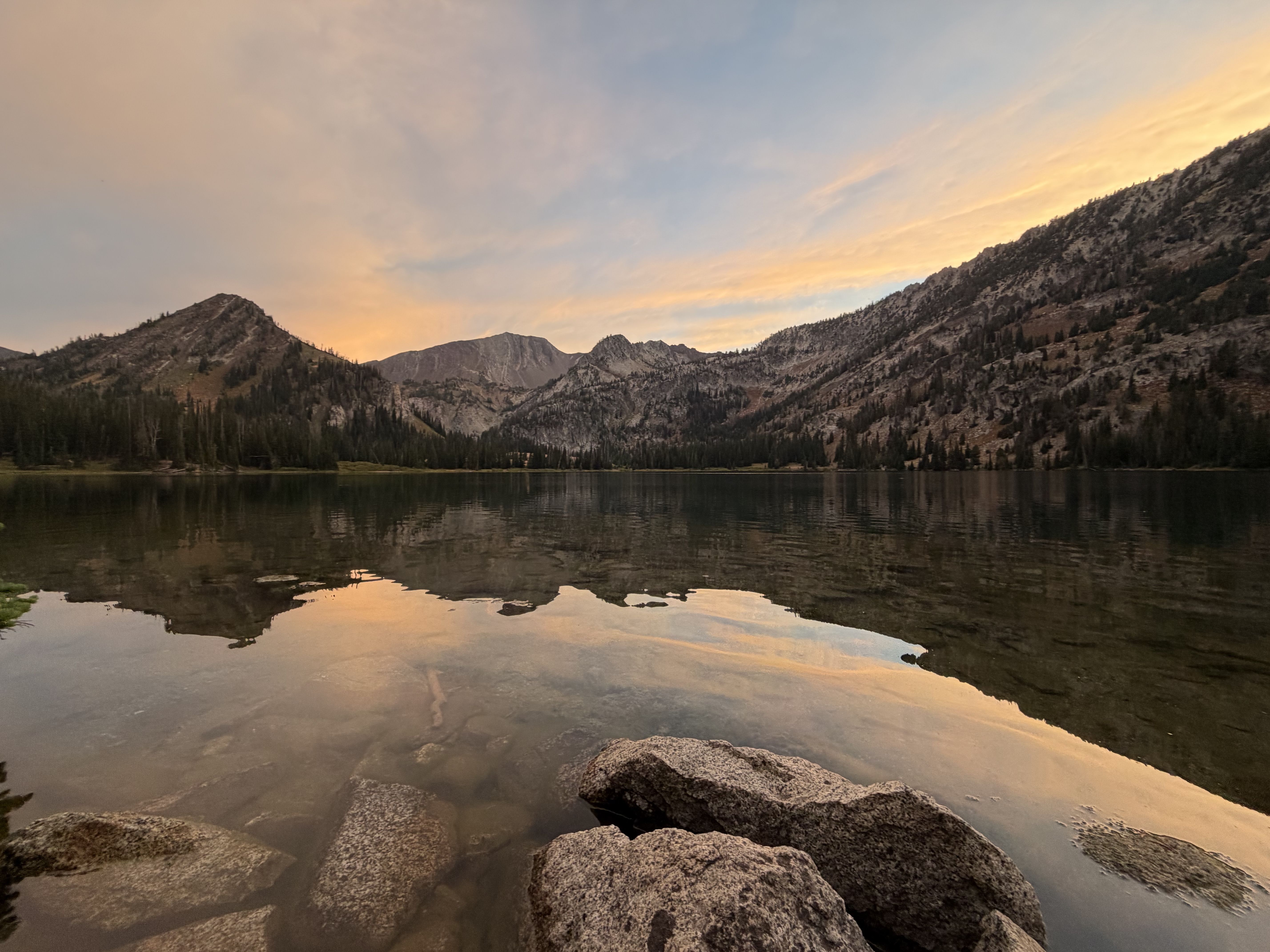 Calm mountain lake at sunset, with jagged forested slopes and a rocky shoreline in the foreground. Warm pink-orange sky reflects on the water.