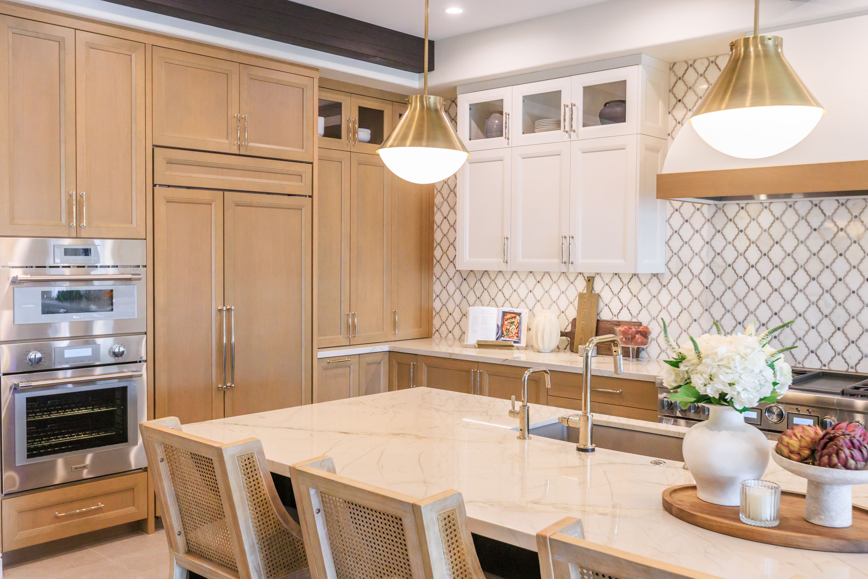 Bright modern kitchen with light wood cabinets on the left and white upper cabinets on the right, a marble island with a double sink, gold pendant lights, stainless ovens, and a white bouquet.