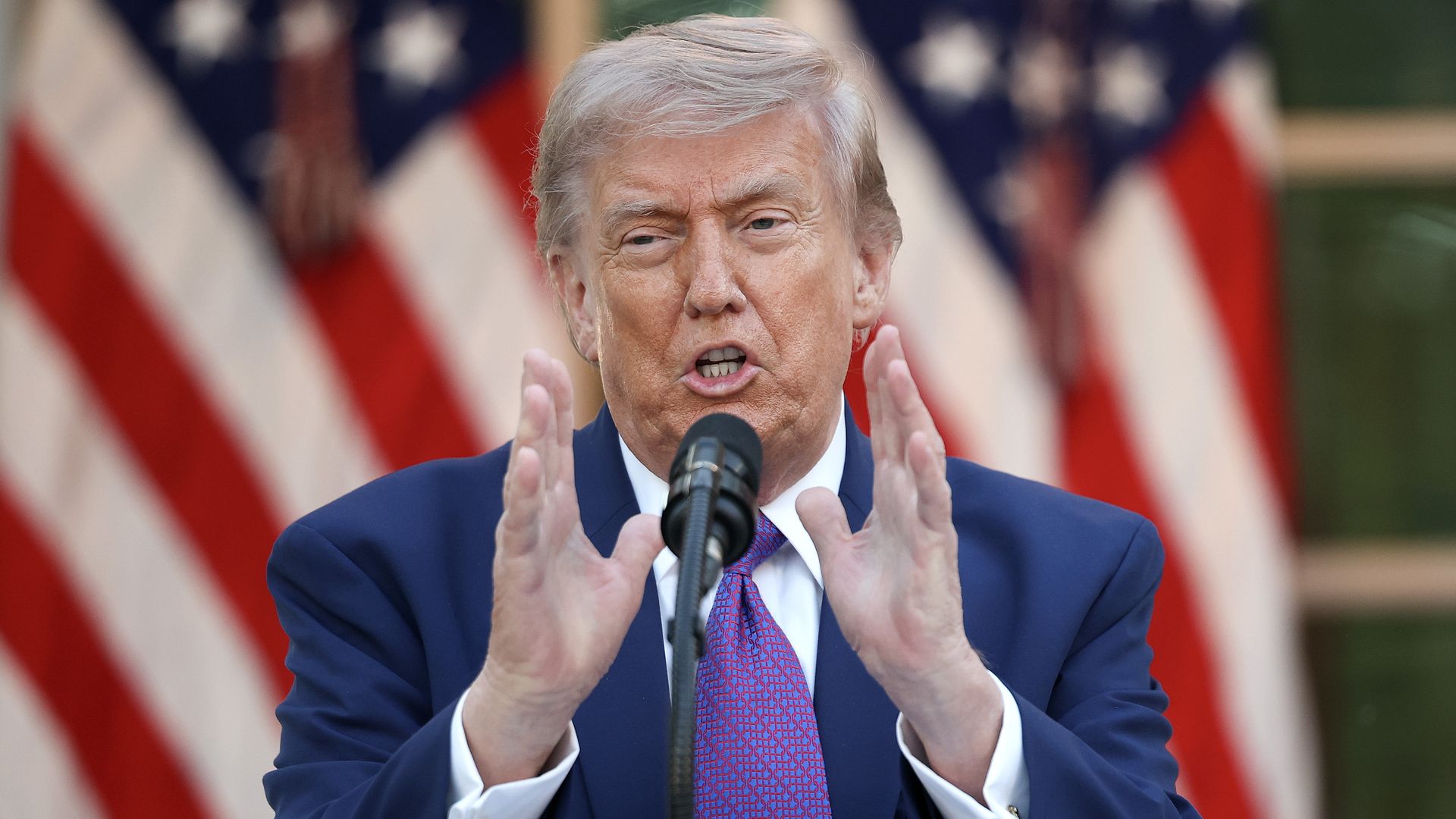 A man in a navy suit and purple tie speaks into a microphone, gesturing with both hands, with blurred American flags in the background.