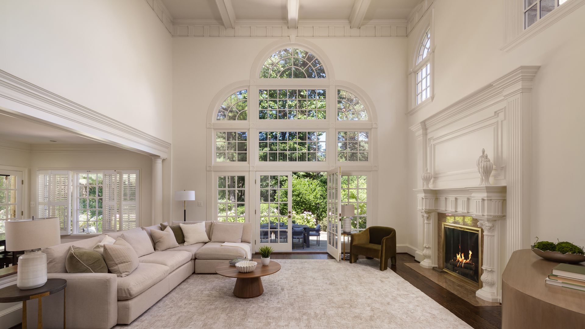 Bright living room with white walls, large arched windows, and French doors to a garden. Beige sectional sofa, round wooden coffee table, and a white ornate fireplace.