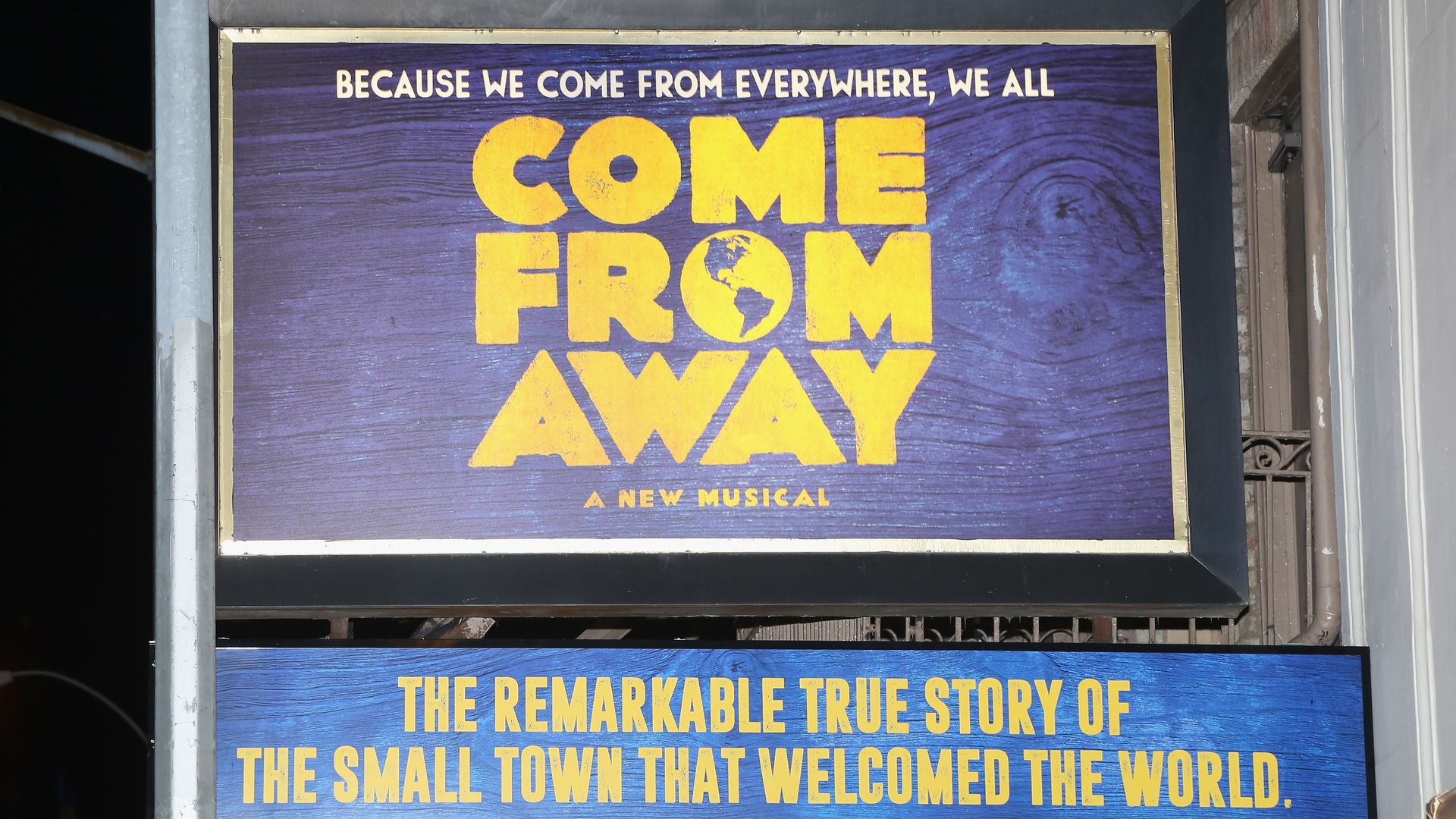 Framed purple poster for the musical "Come From Away" with huge yellow letters 'COME FROM AWAY' and a globe in the O; top line reads "BECAUSE WE COME FROM EVERYWHERE, WE ALL".
