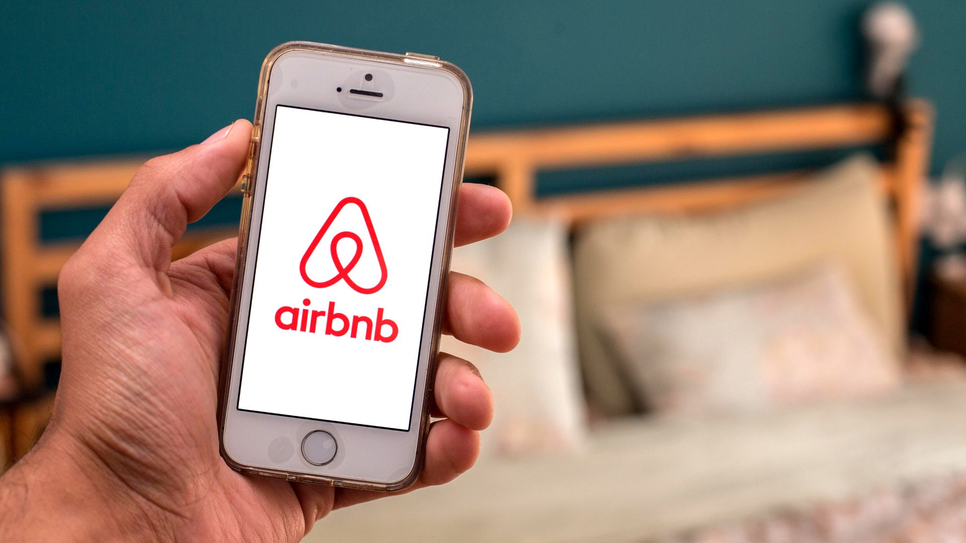 Photo illustration of the Airbnb logo on a smartphone with a bed in the background