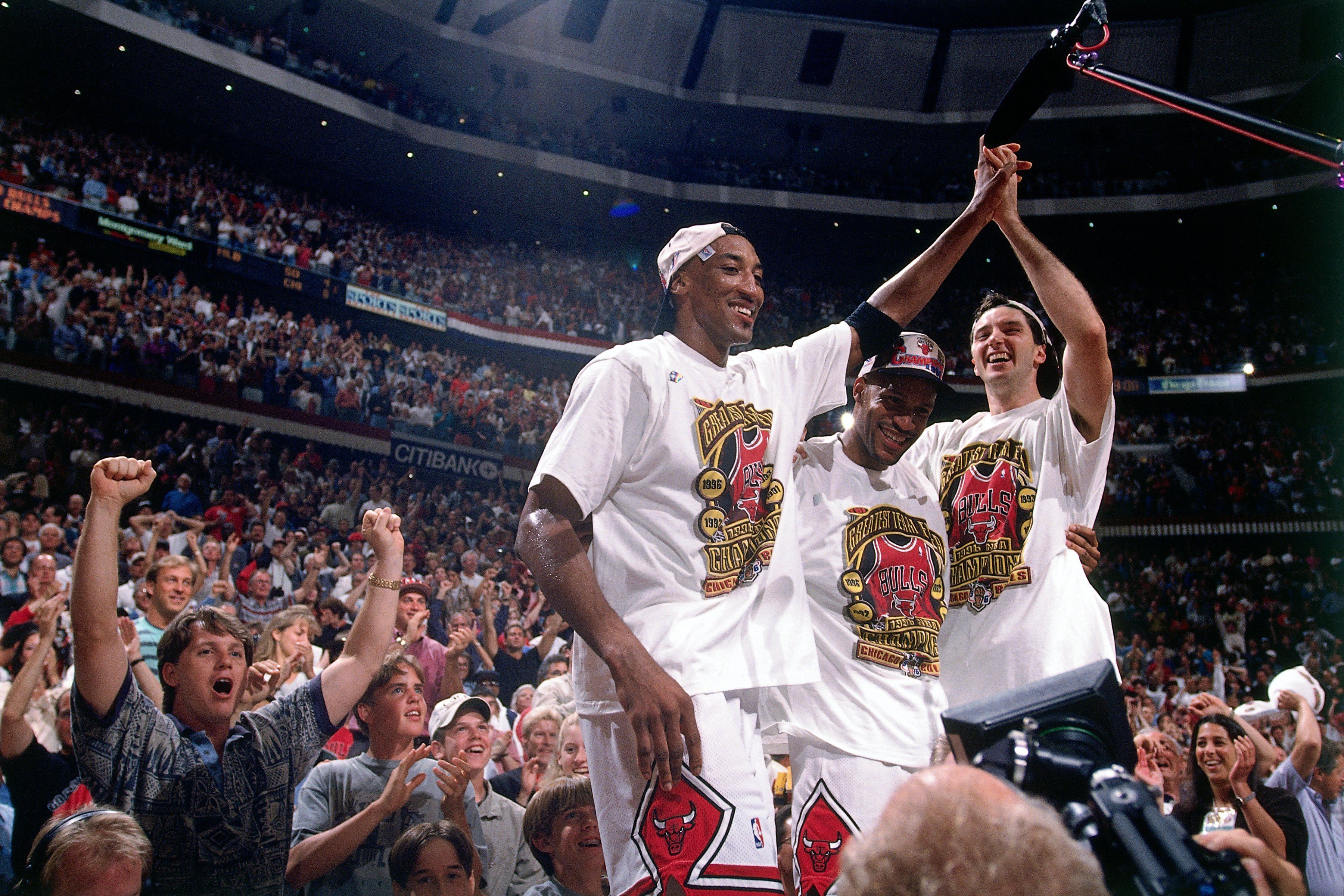 Three Chicago Bulls players celebrate on court, high-fiving with white shirts featuring Bulls logos; a packed arena cheers in the background.
