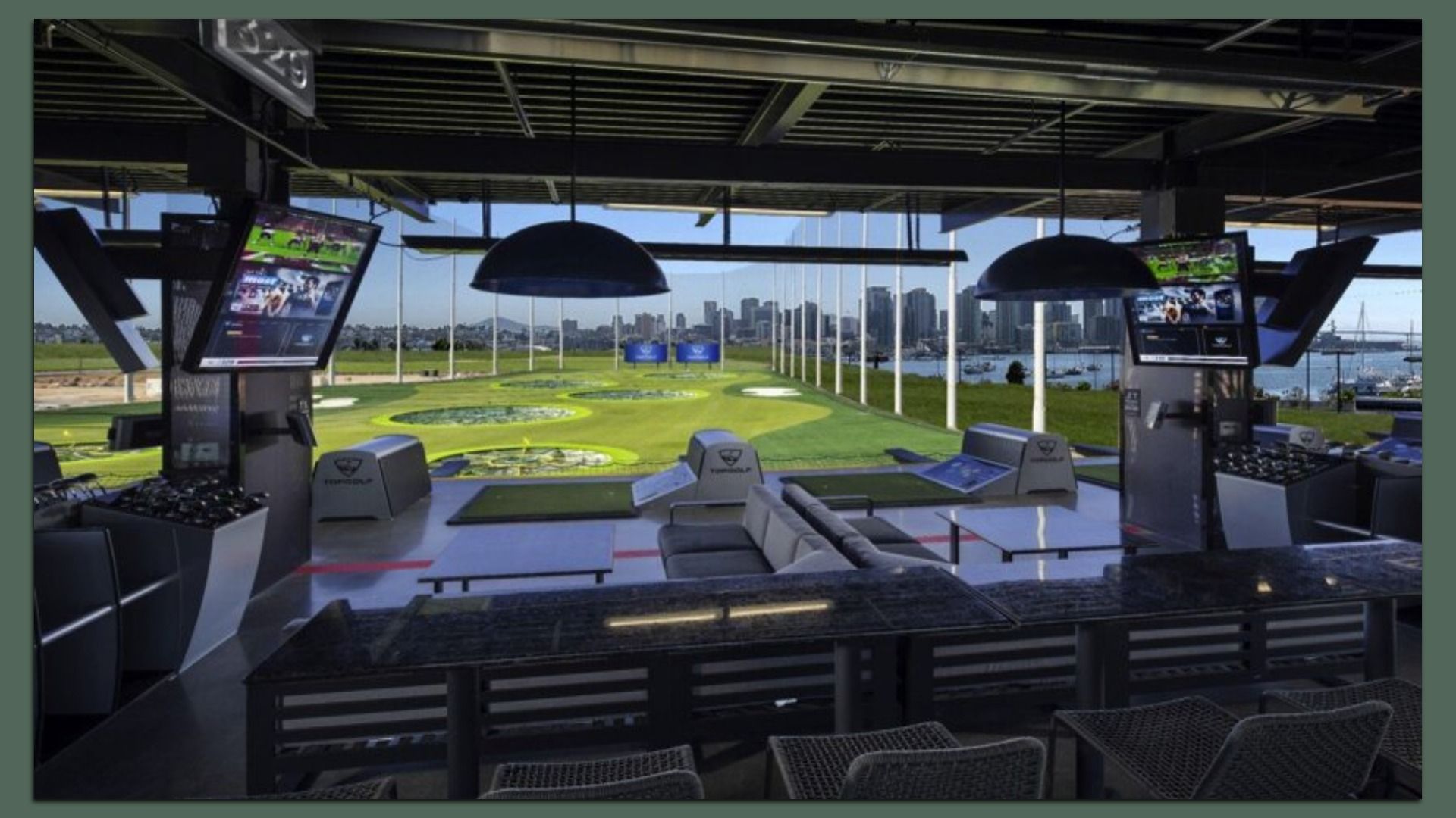 Indoor golf driving range bay with two large screens on each side, black ceiling beams, and green turf with circular target zones. A city skyline by water is visible through the glass windows.