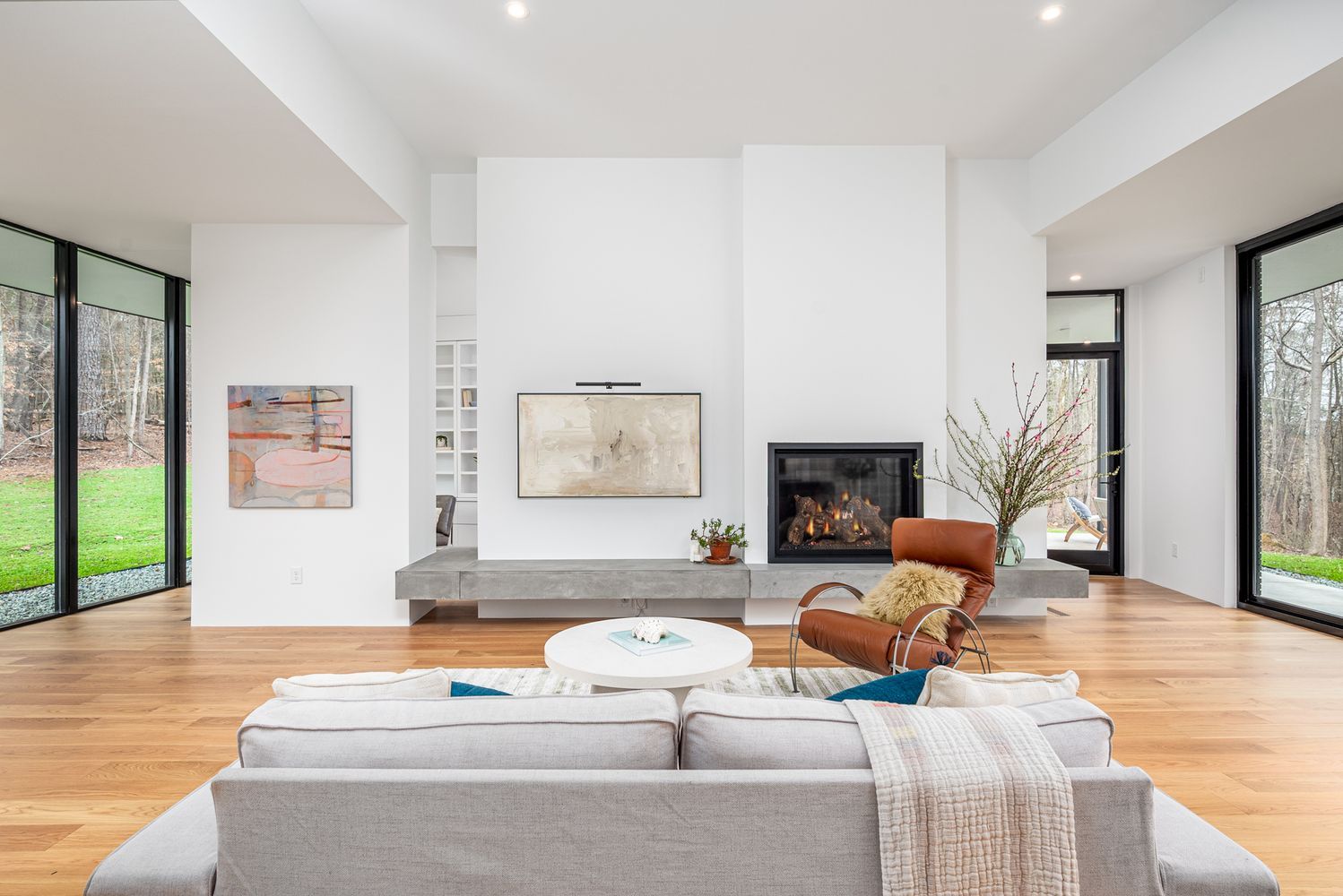 Modern living room with light gray sofa, round white coffee table, brown leather chair with yellow pillow, fireplace, abstract paintings, and large windows showing green lawn and trees outside.